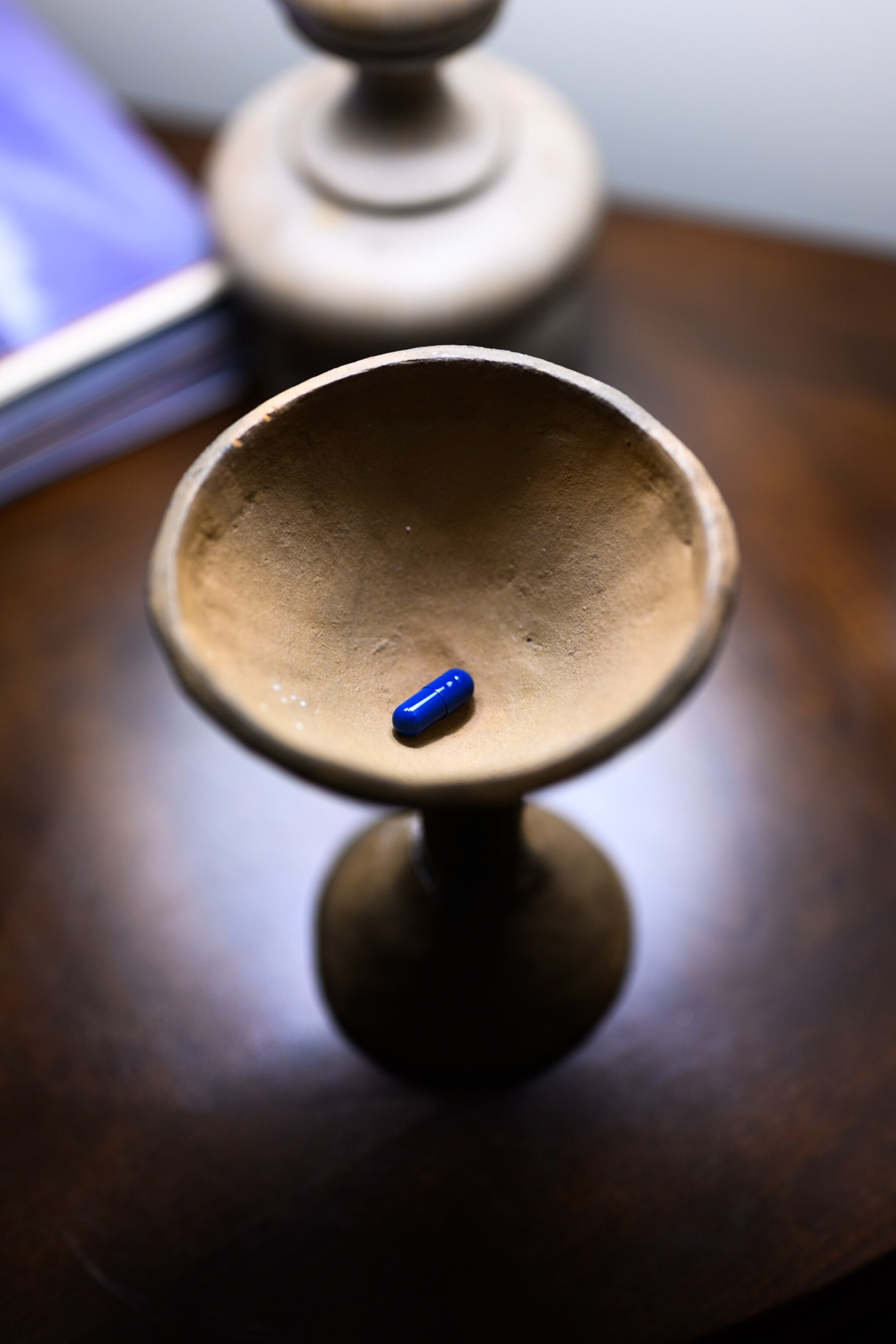 A single blue pill capsule, containing a dose of psilocybin, is displayed in a ceramic chalice on a wooden table.