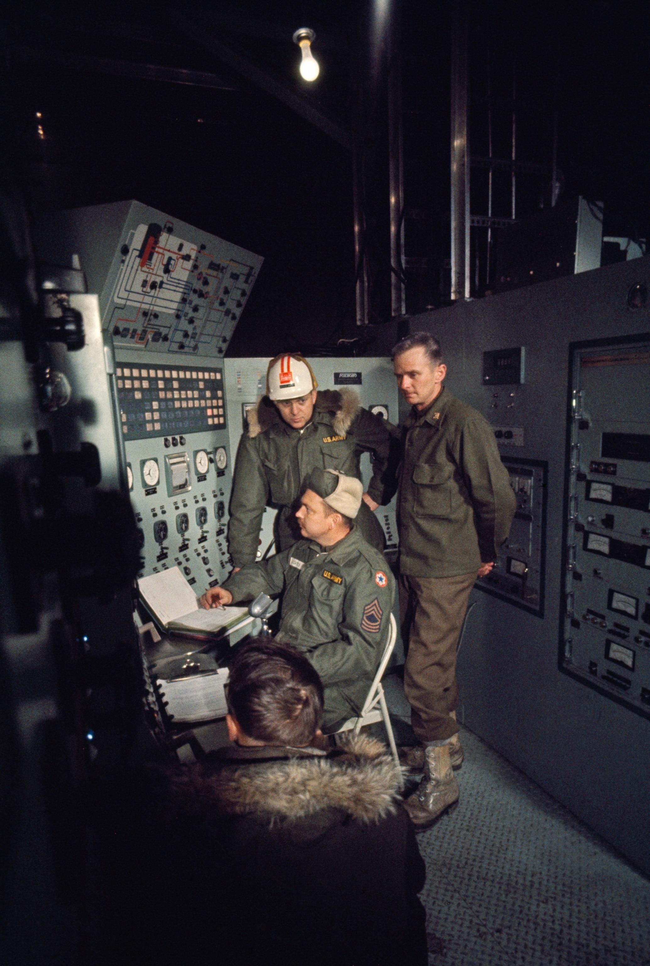 Four specialists are photographed monitoring a control pannel, with one of the men writing in a book.