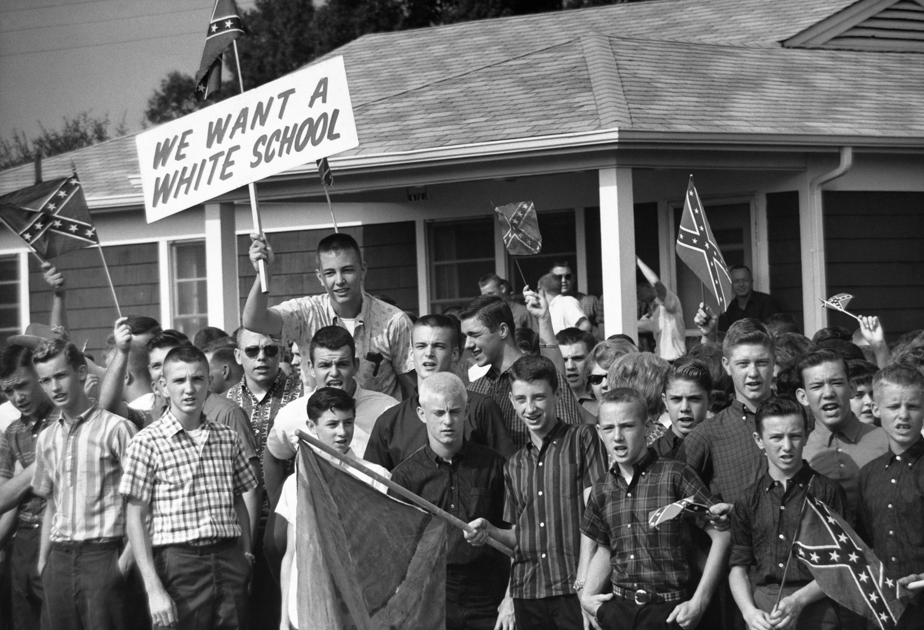 A crowd of White boys hold confederate flags and a sign advocating for a white school