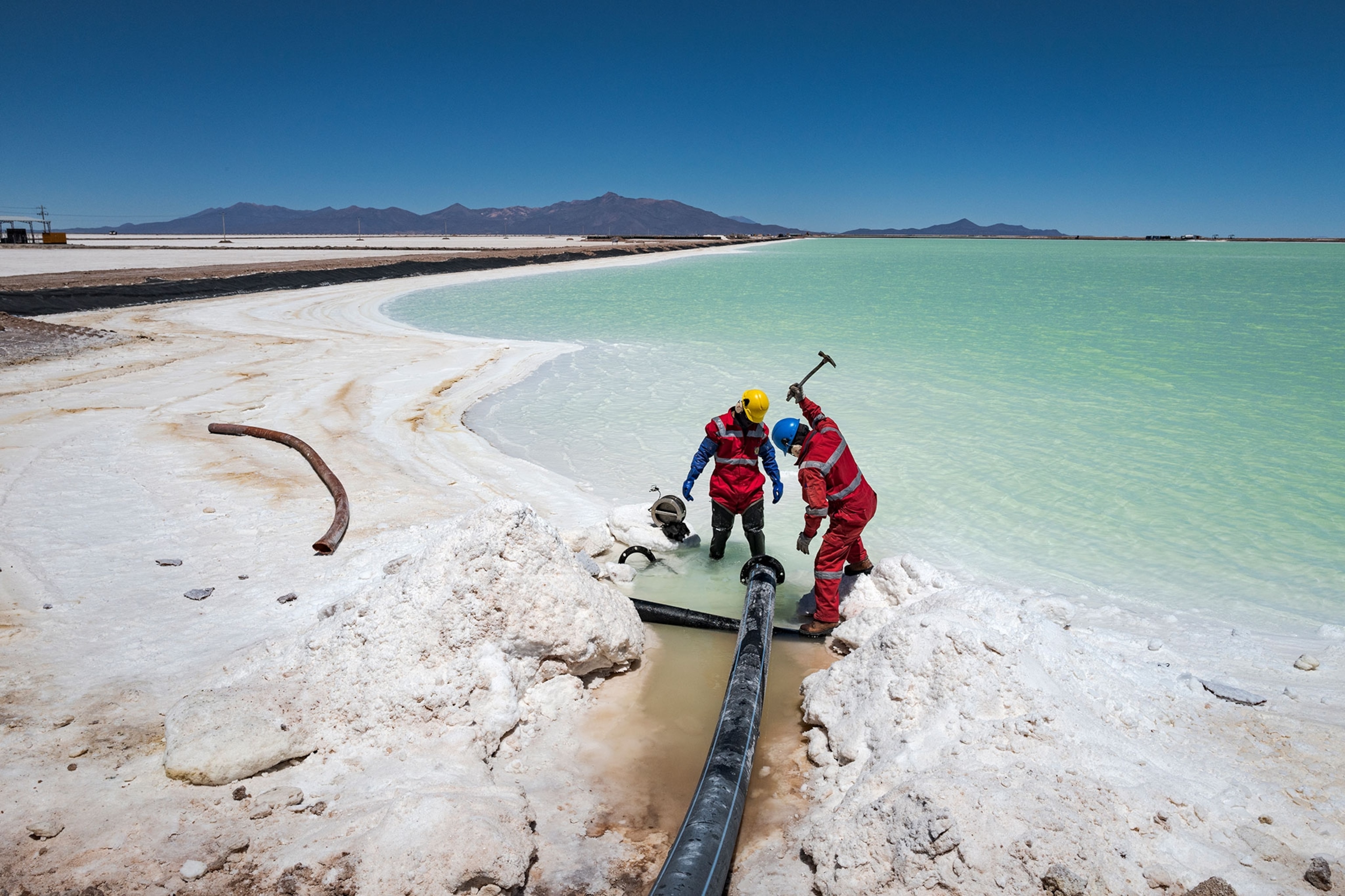 Workers, wearing red, one piece suits, at the lithium pilot plant use hammers to break up a layer of salt that periodically clogs the pipes transporting brine with lithium into large, turquoise blue evaporation pools.