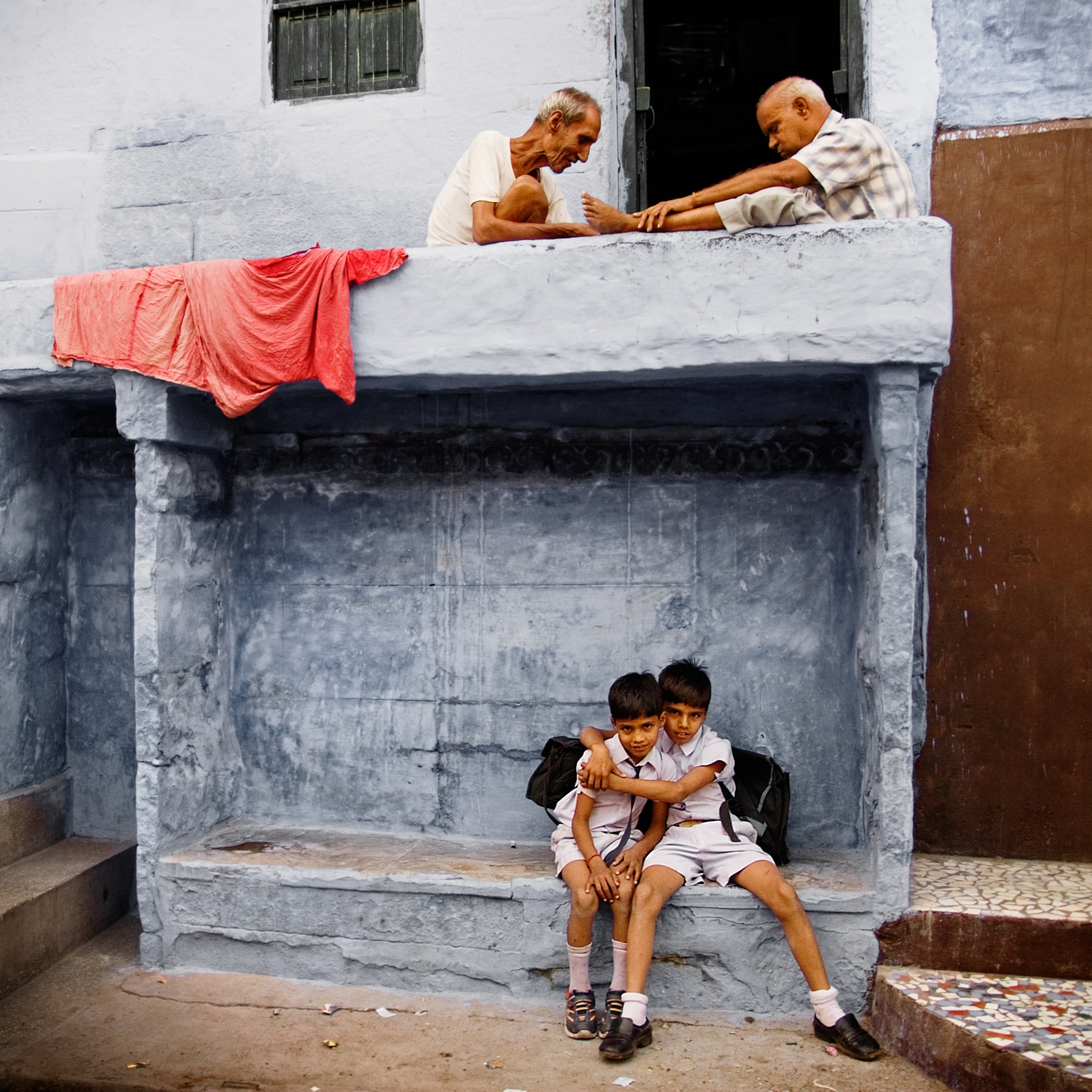 Two young boys embracing and looking at the camera while seated beneath a concrete balcony, upon which two elderly men are playing chess.