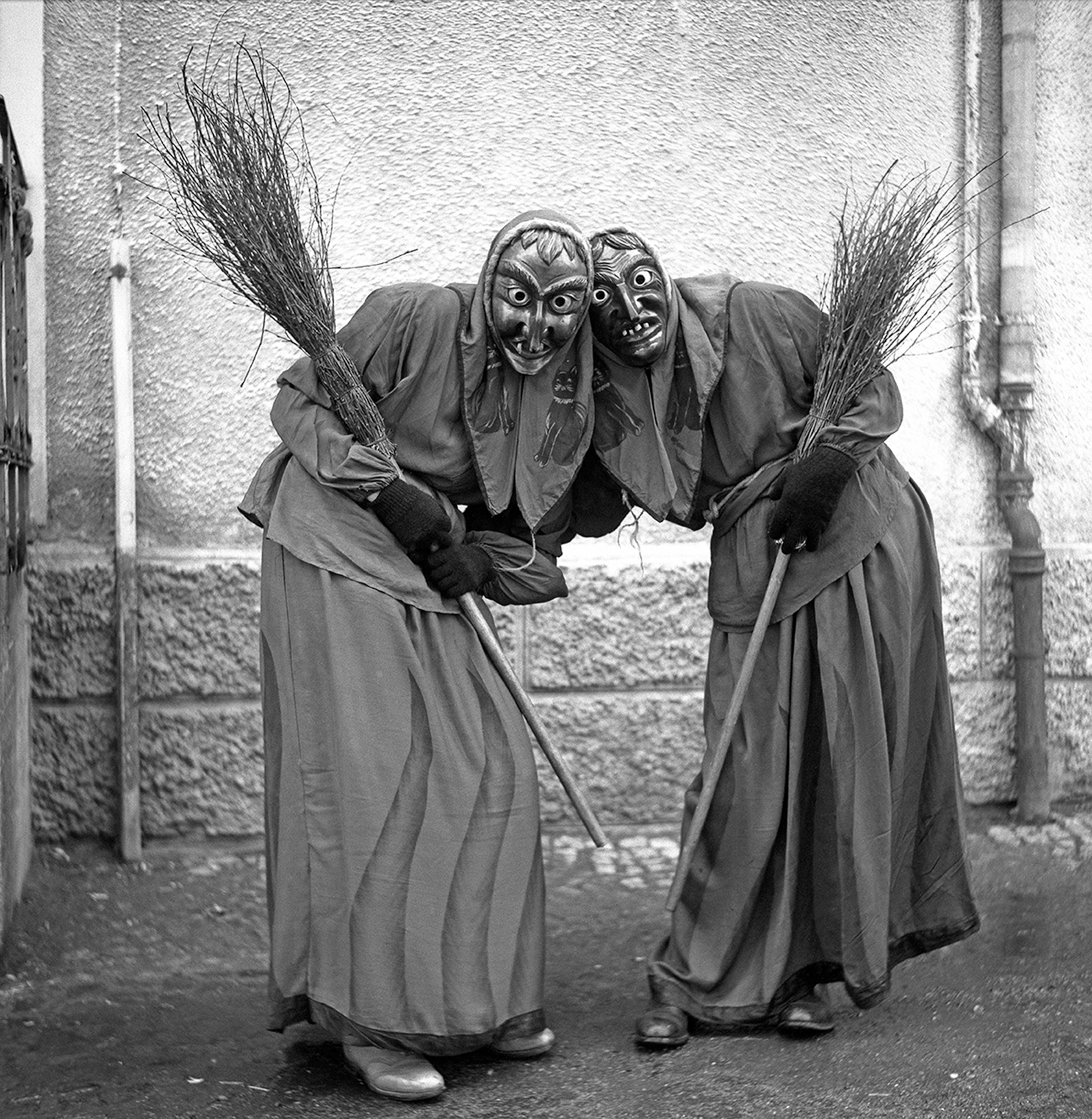 Two people in witch costumes with scary masks lean toward each other. They both have brooms. The photo is black and white.
