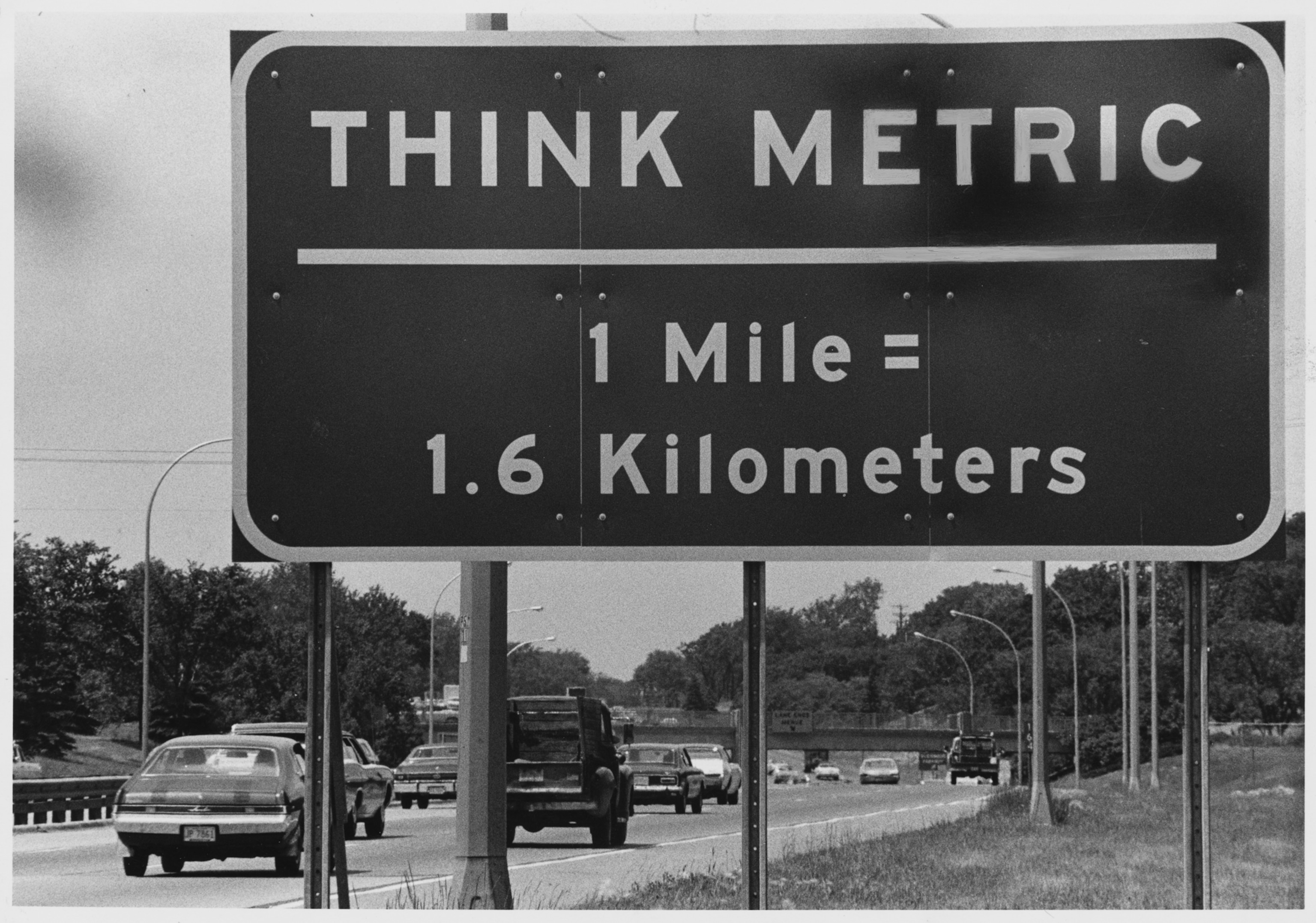 A traffic sign near the US border in Quebec reads "THINK METRIC, 1 Mile = 1.6 Kilometers."