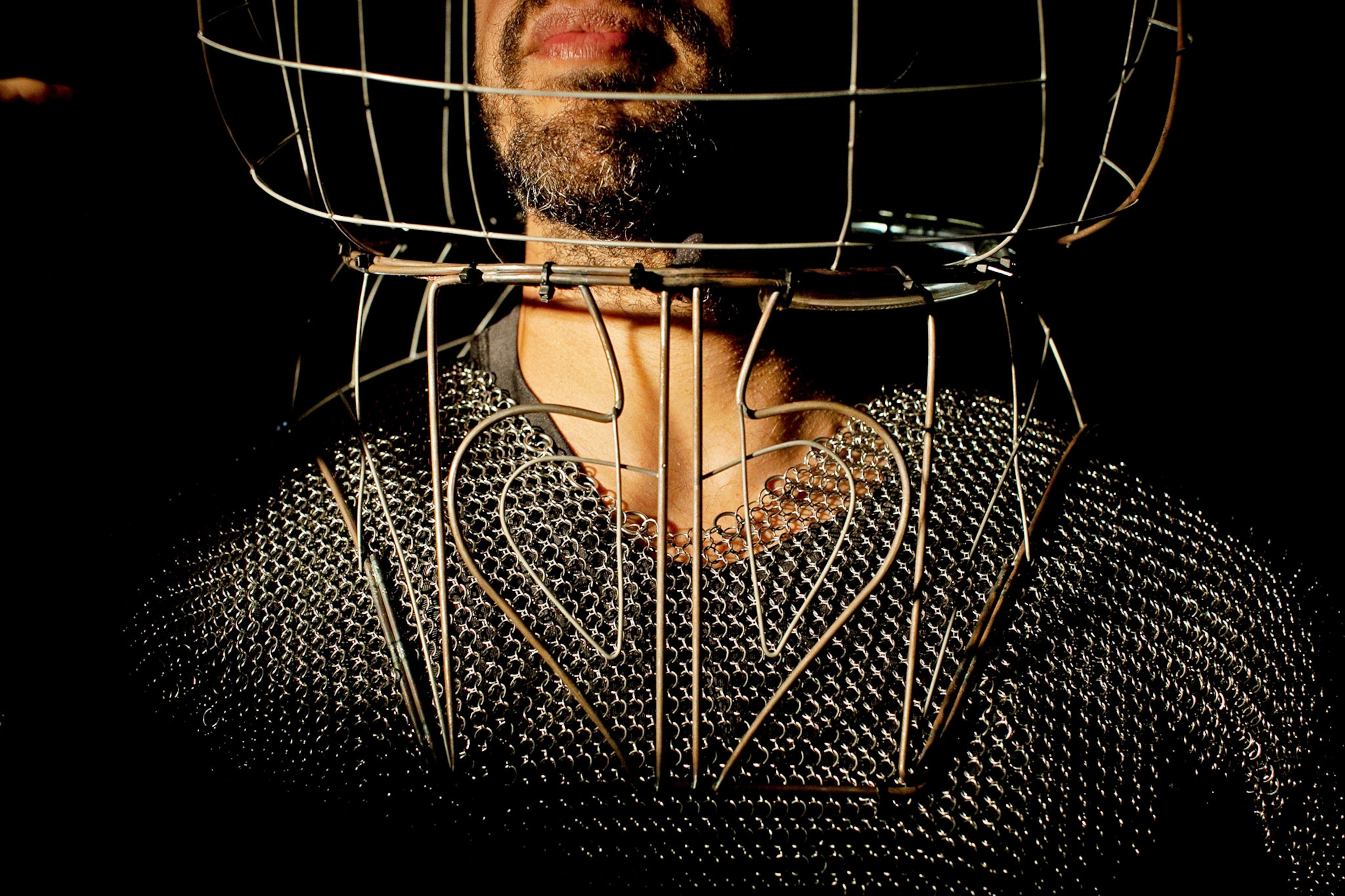 A close-up view of a man wearing a metal suit similar to chain mail, with a cage-like metal helmet
