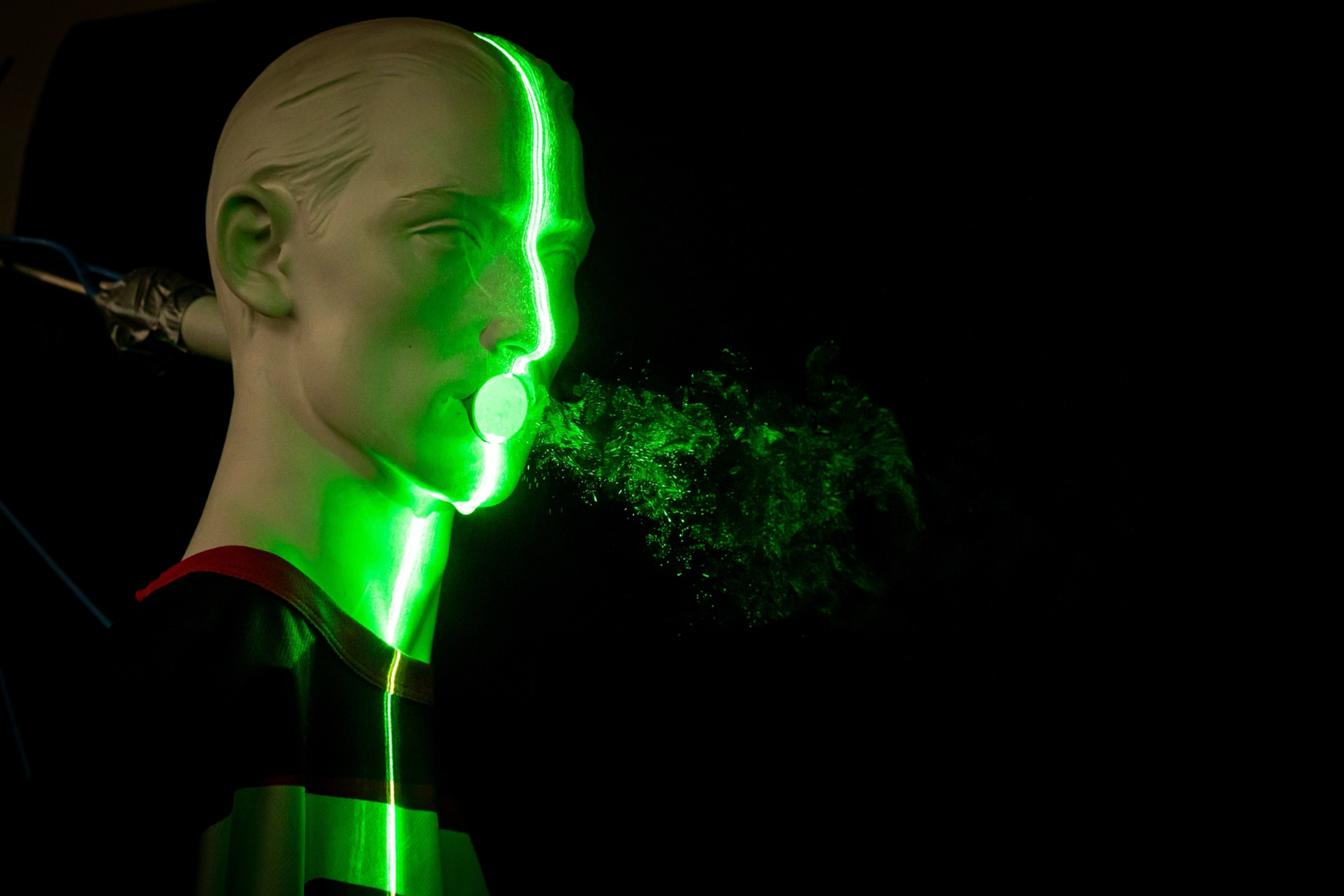 A mannequin head spewing droplets from its mouth in a lab simulated cough, lit by a beam of green light