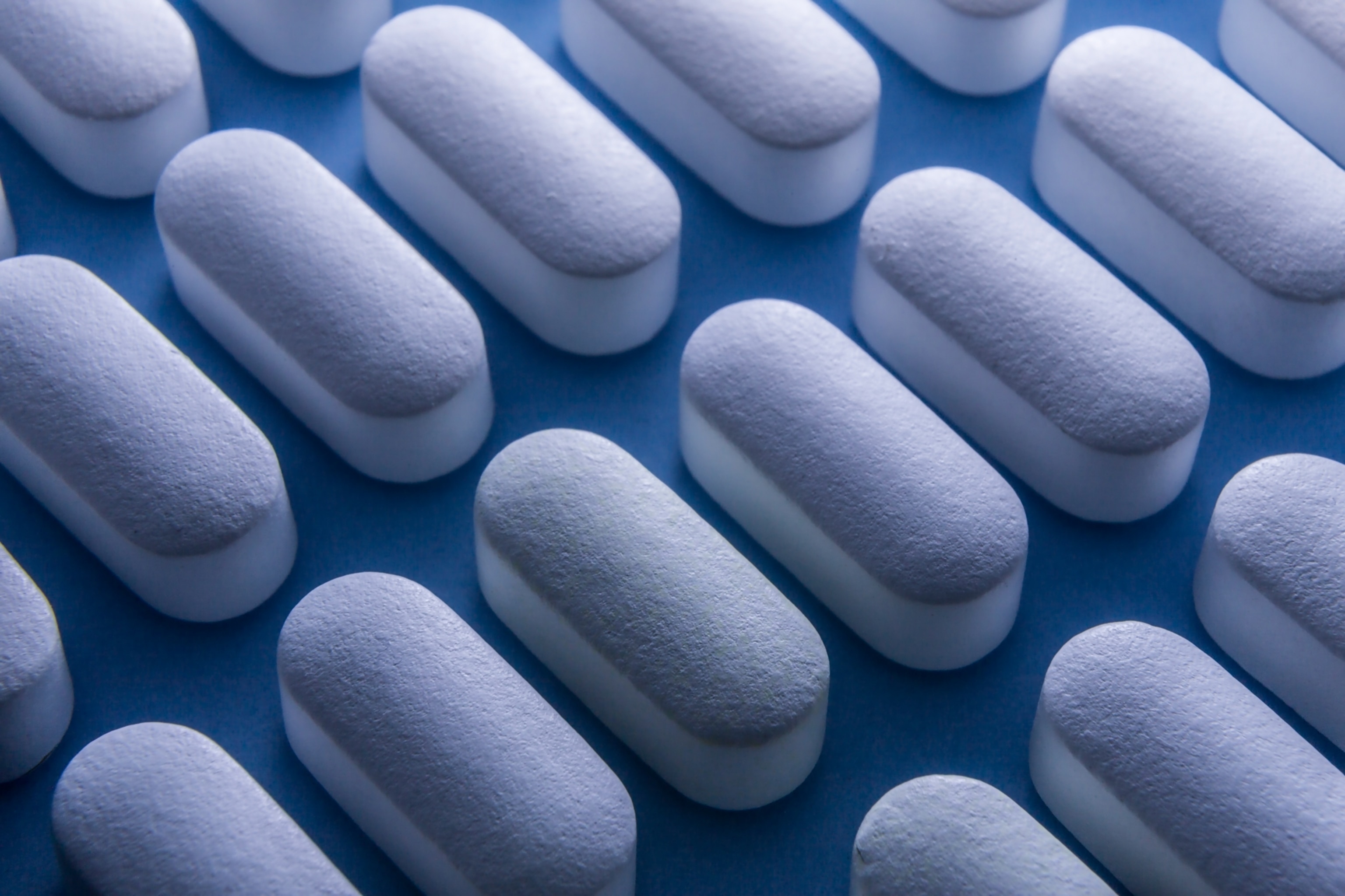 A close-up view of rows of generic white pills lined diagonally across the frame, against a blue background.