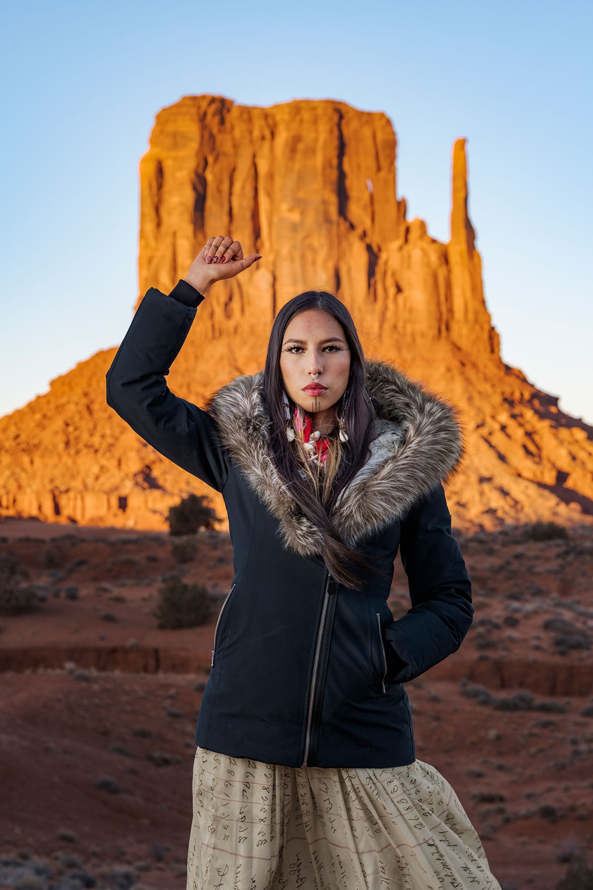 Portrait of a female, Navajo model, wearing a black coat with a fur trimmed hood, standing with her fist raised in front of sacred land in Monument Valley.