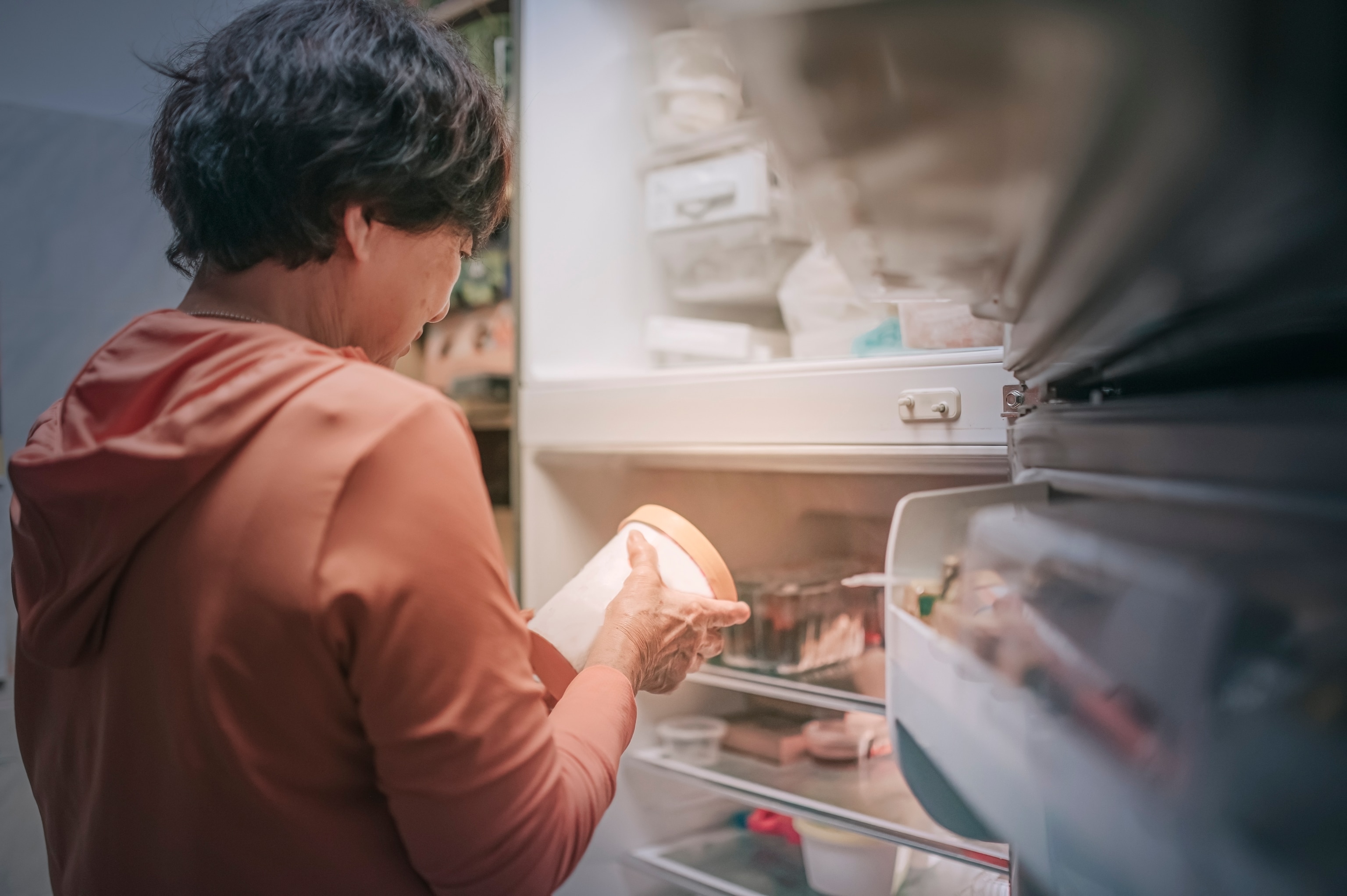 Side view of a senior Asian woman holding a food container in front of an open refrigerator.