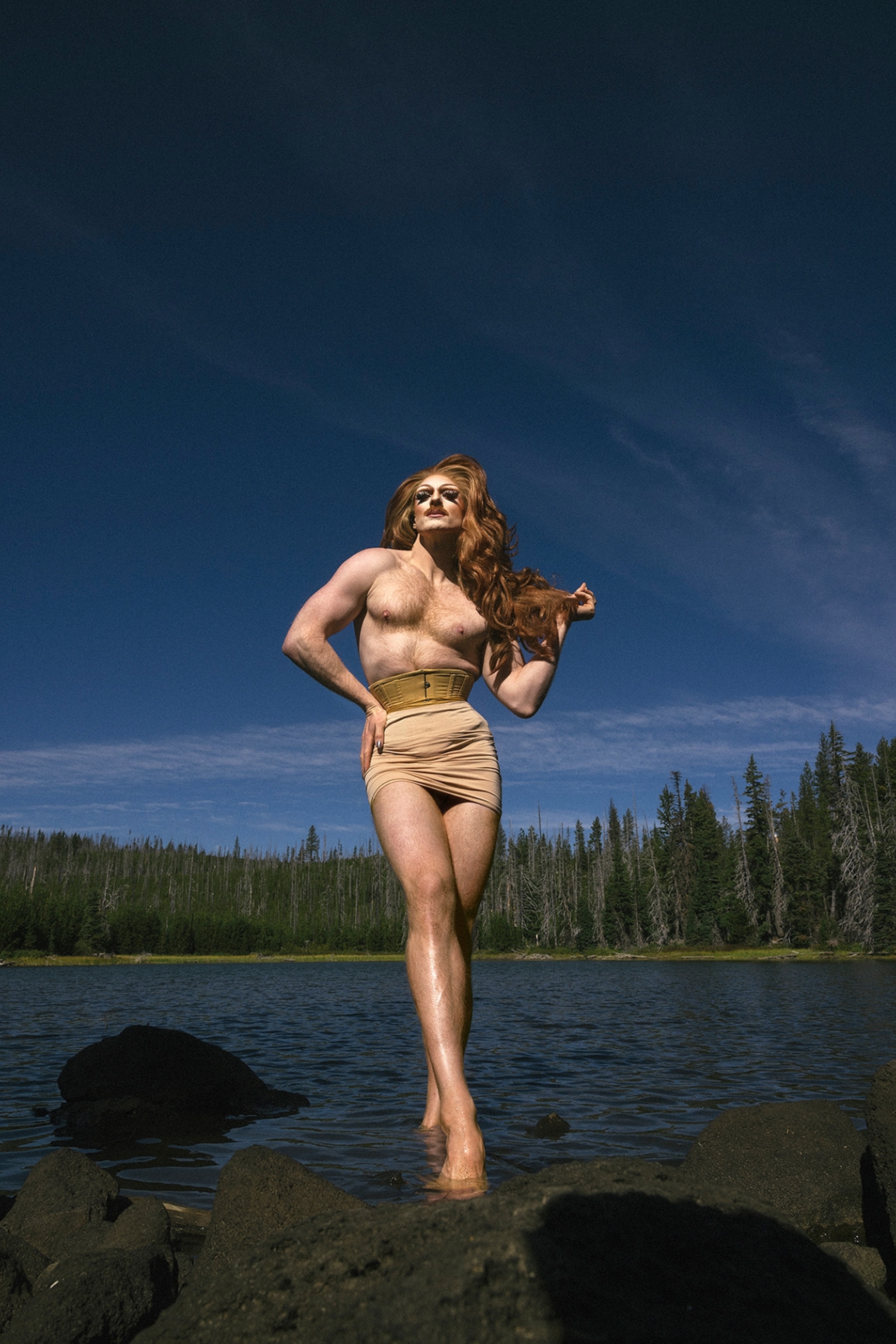 A drag queen with long wavy brown hair wearing only a small corset and skirt stands posing by a lake with pine trees lined in the background and a blue sky above