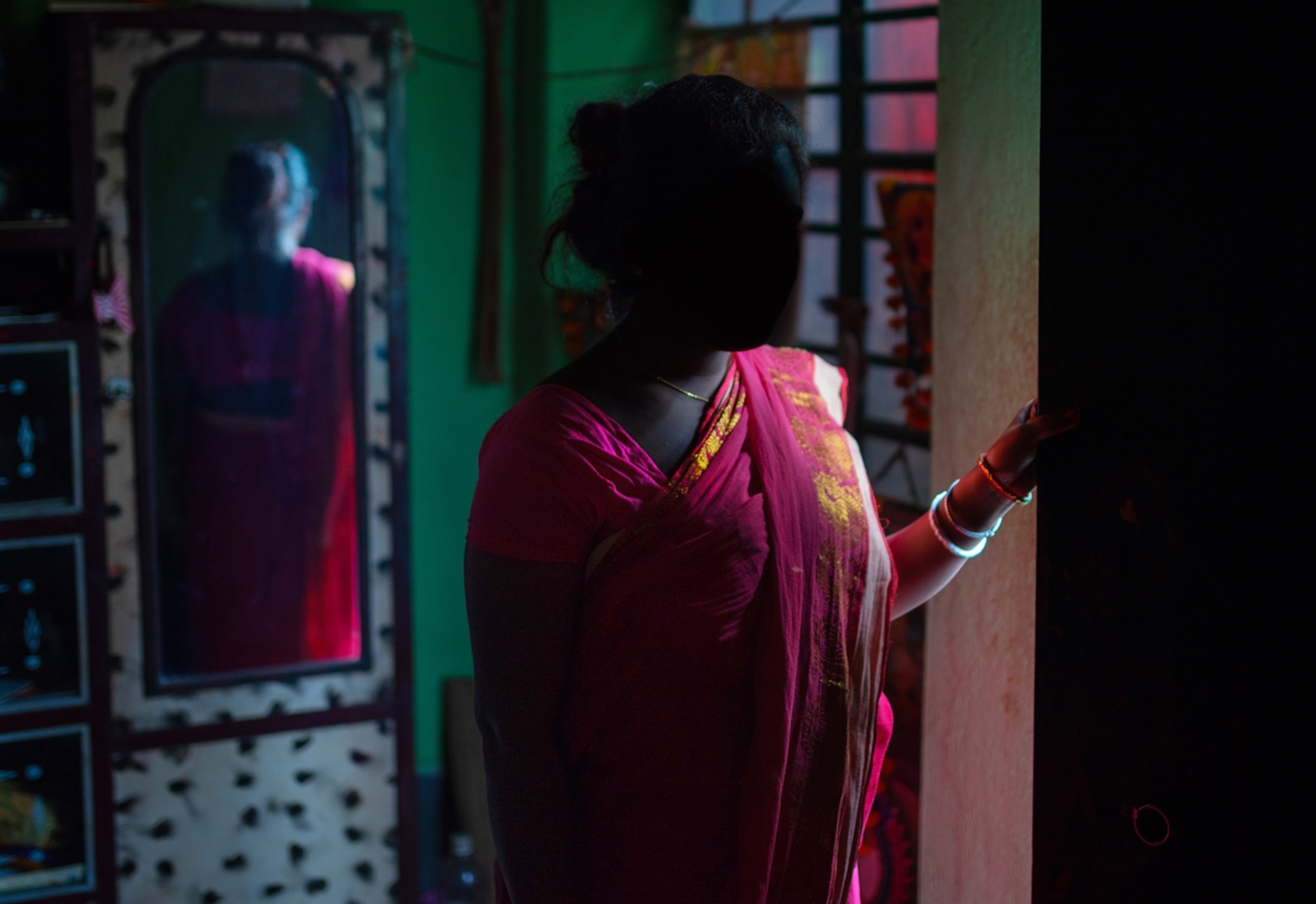 a woman wearing a pink dress, silhouetted by the light of the room, peers around a wall