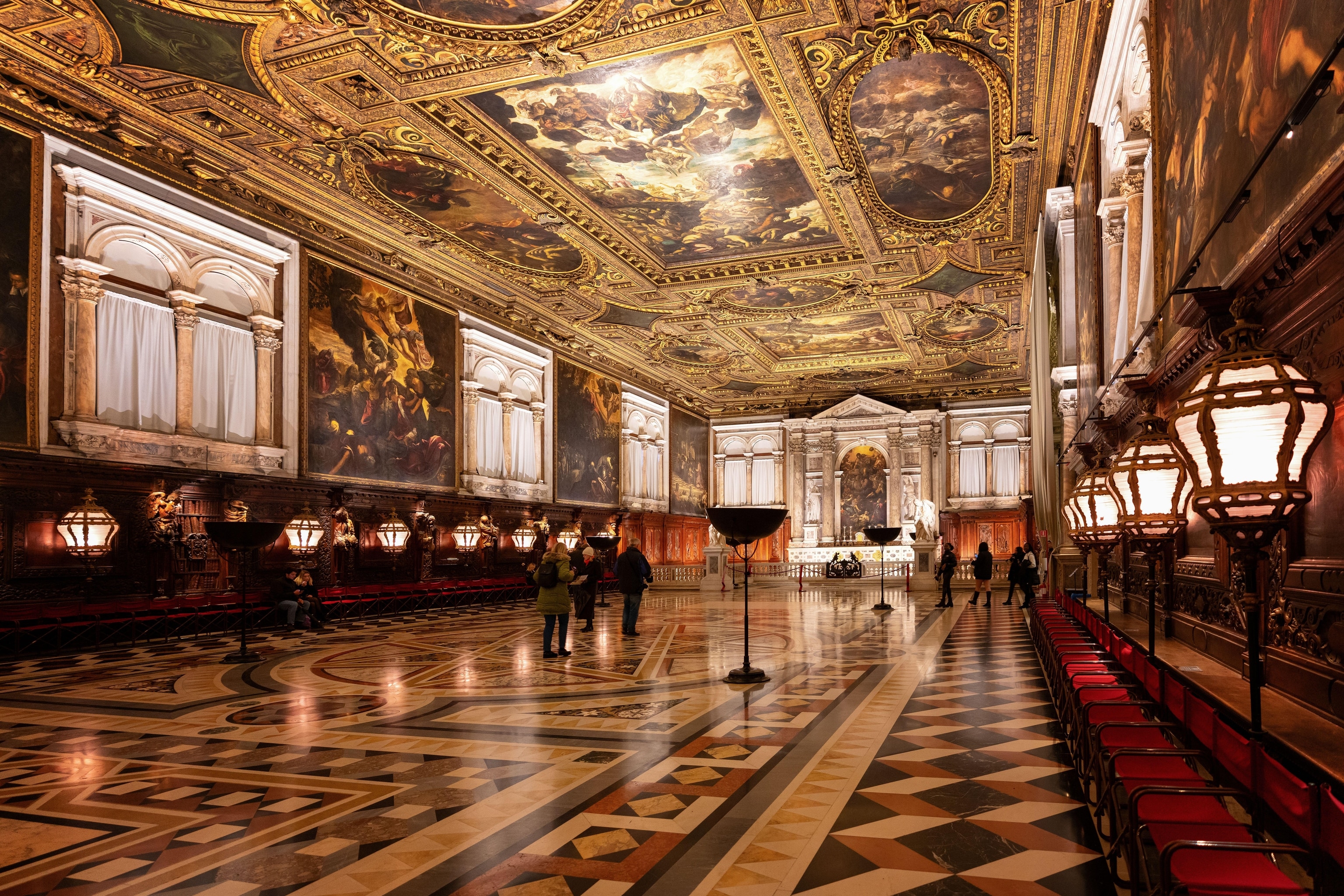 The interiors of an old building in Venice. The ceiling is gilded in gold and has paintings on the ceiling. The floors are tiled in a mosaic pattern.