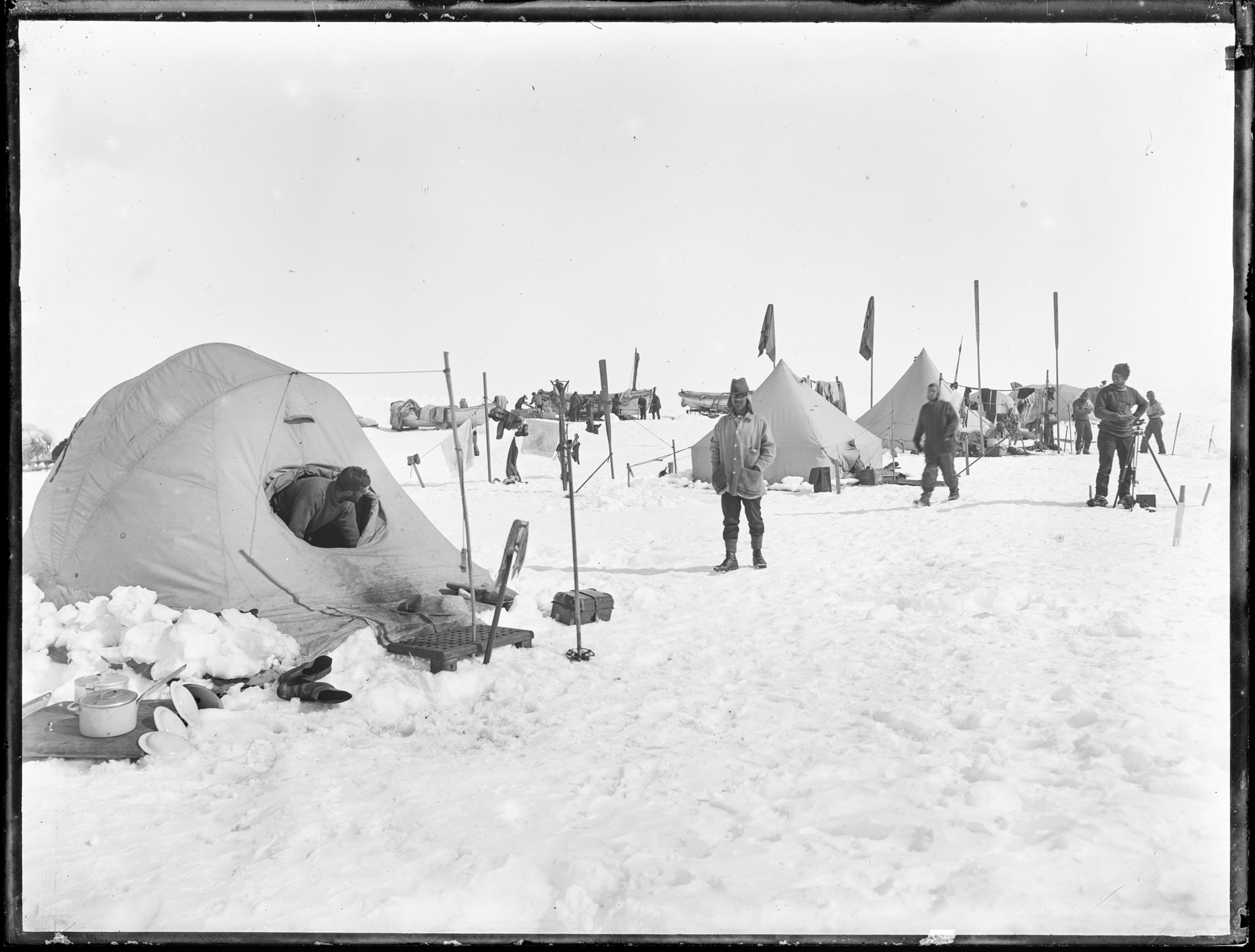 Photograph of the camp set up in the snow. There are tents with pots, plates, and boots outside