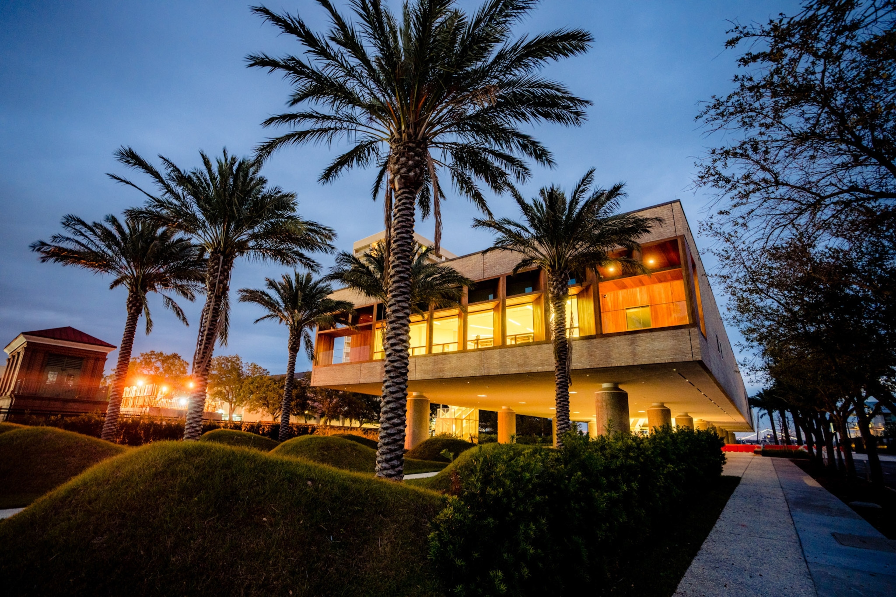 The warm lights of the f International African American Museum illuminate the interior with trees in foreground at night.