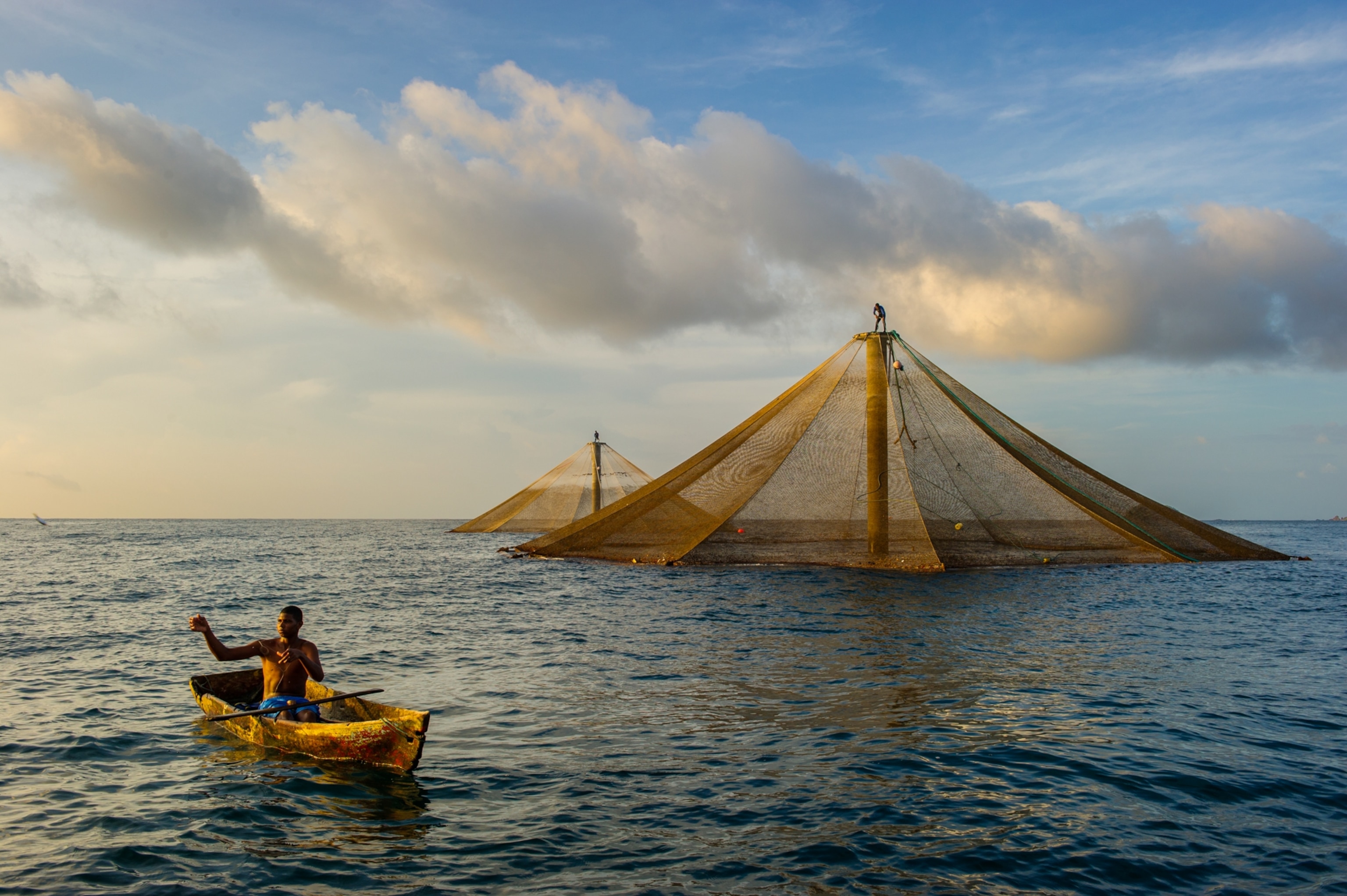 A man seated in a small rowboat extends his arms to catch an object. Behind him, two diamond-shaped fish cages rise from the water.