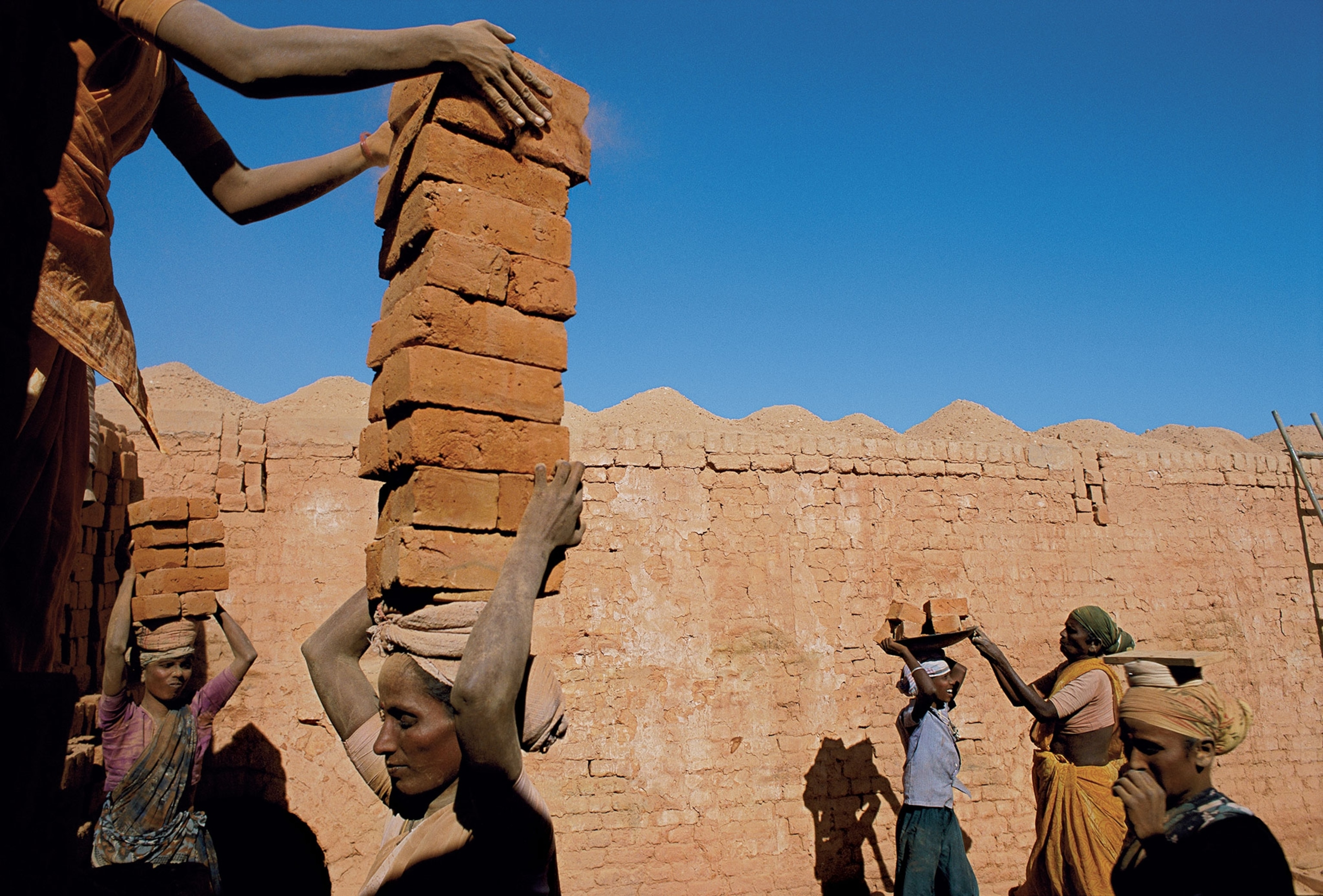 Several people carrying bricks on their heads in front of a brick wall and blue sky
