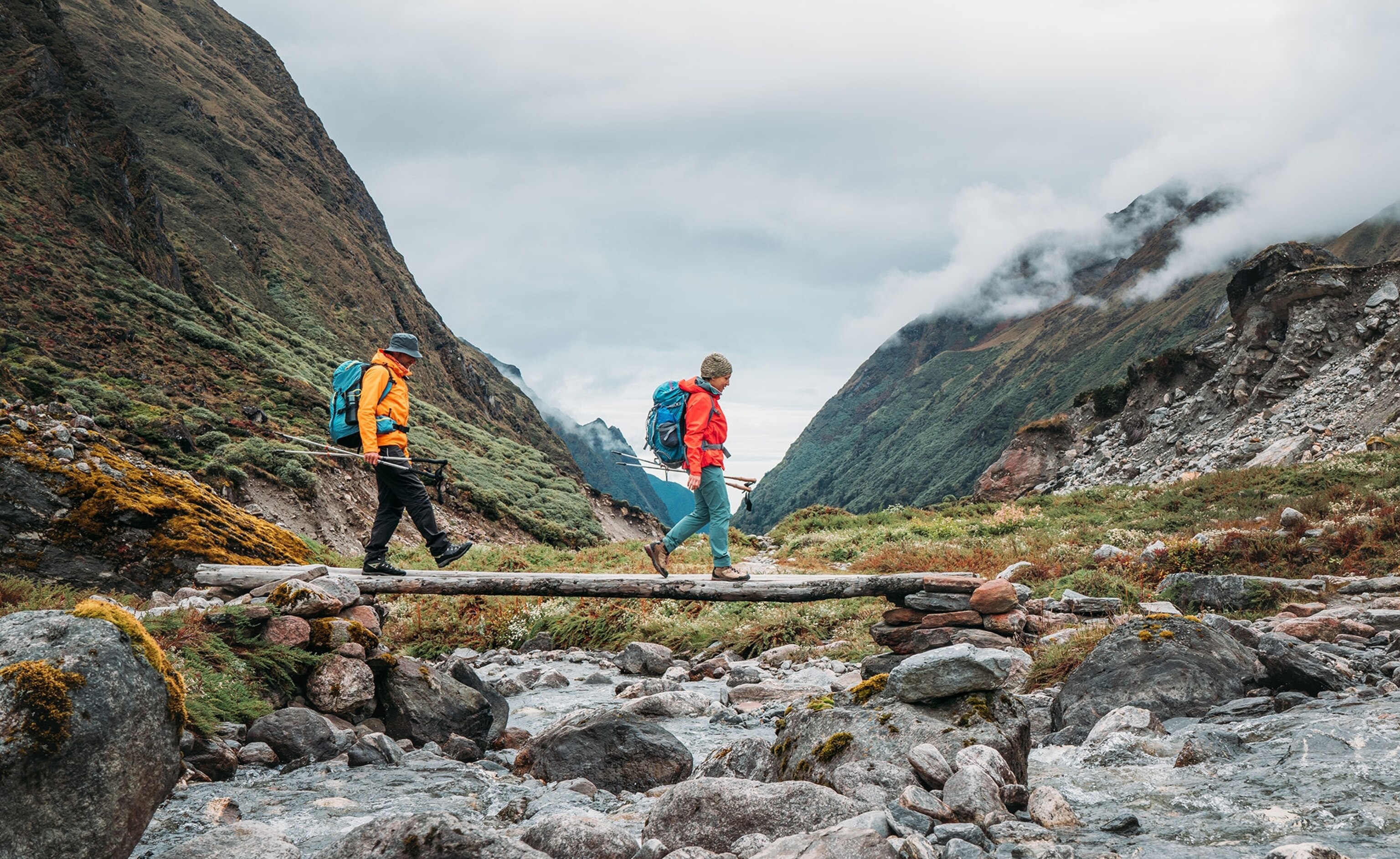 Two people walking across wood log over a stream of water.