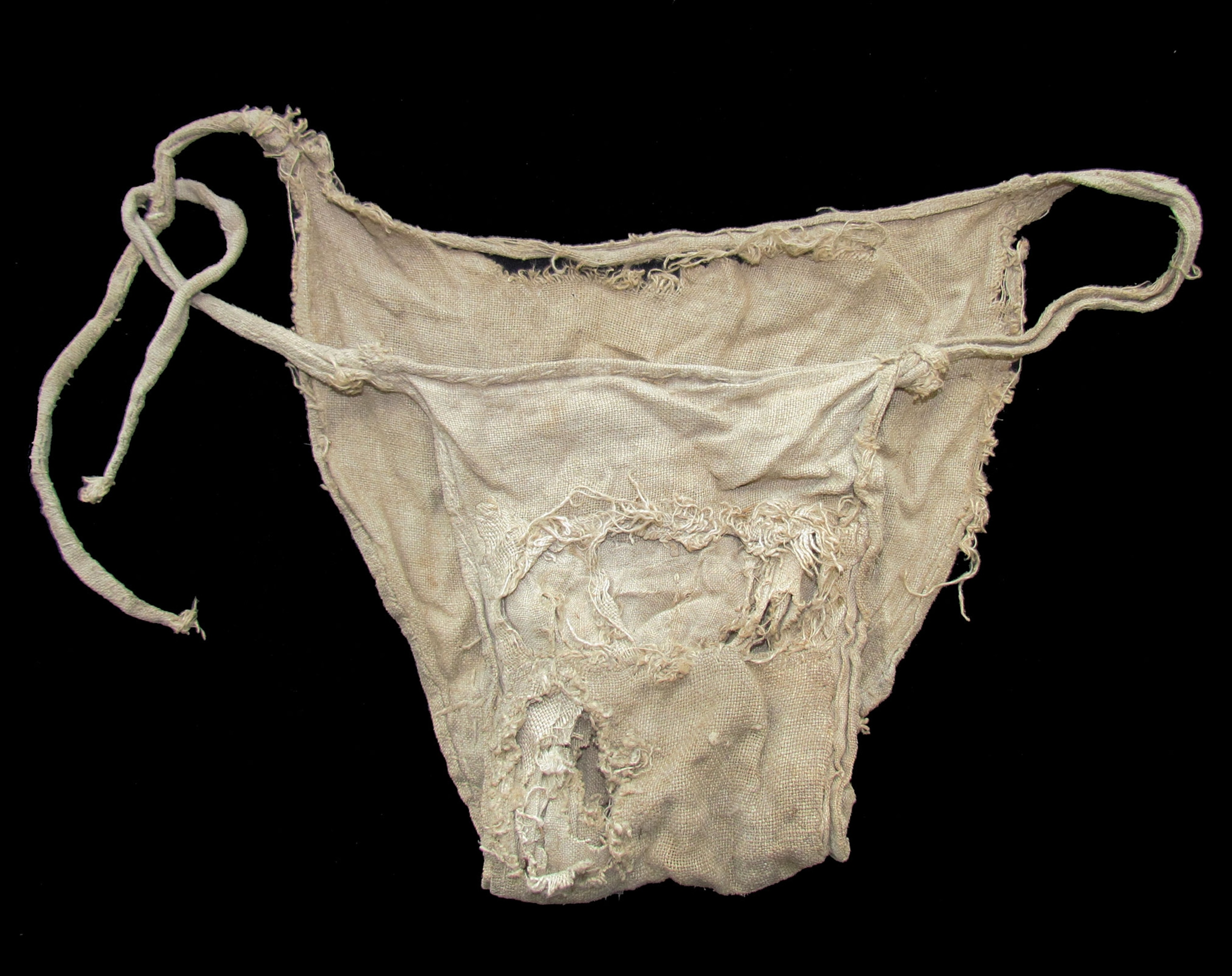 A whitish/brown, torn, triangular cloth with long, white strings connecting the front part of the cloth to the back part of the cloth against a black background.