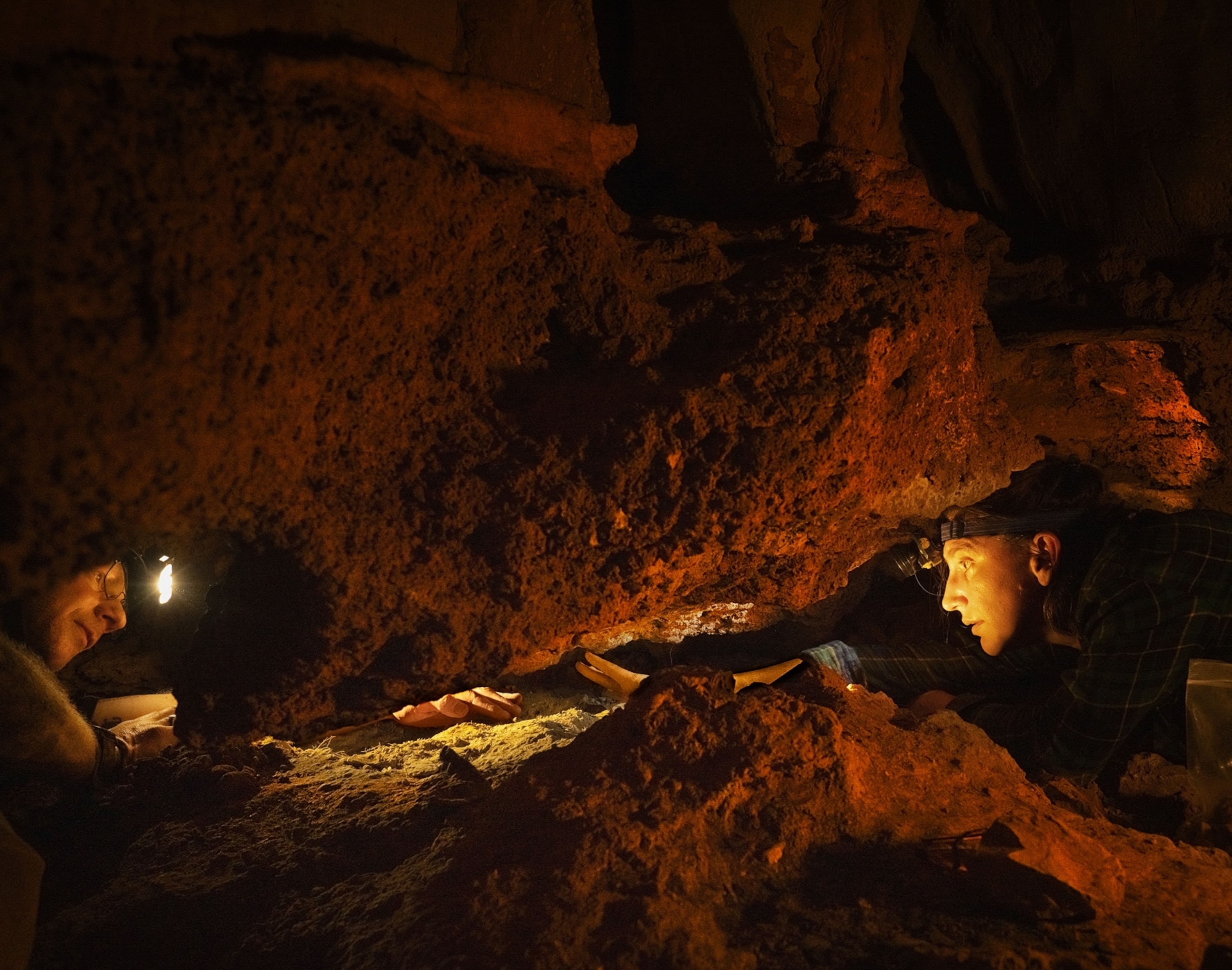 Two people try to reach into a small nook within a cave.