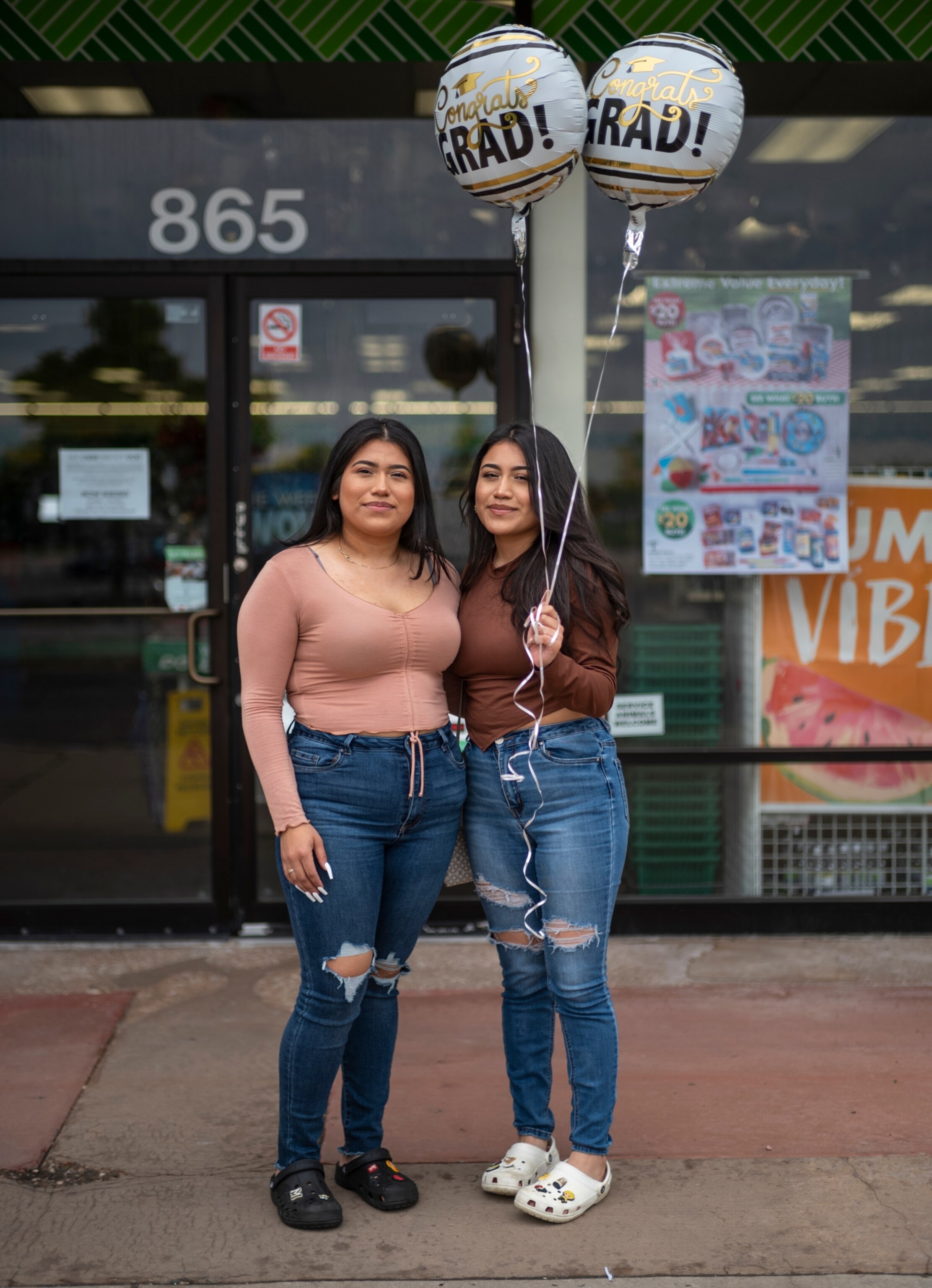 two teenaged women standing in front of a store holding two mylar balloons that say Grad on them.