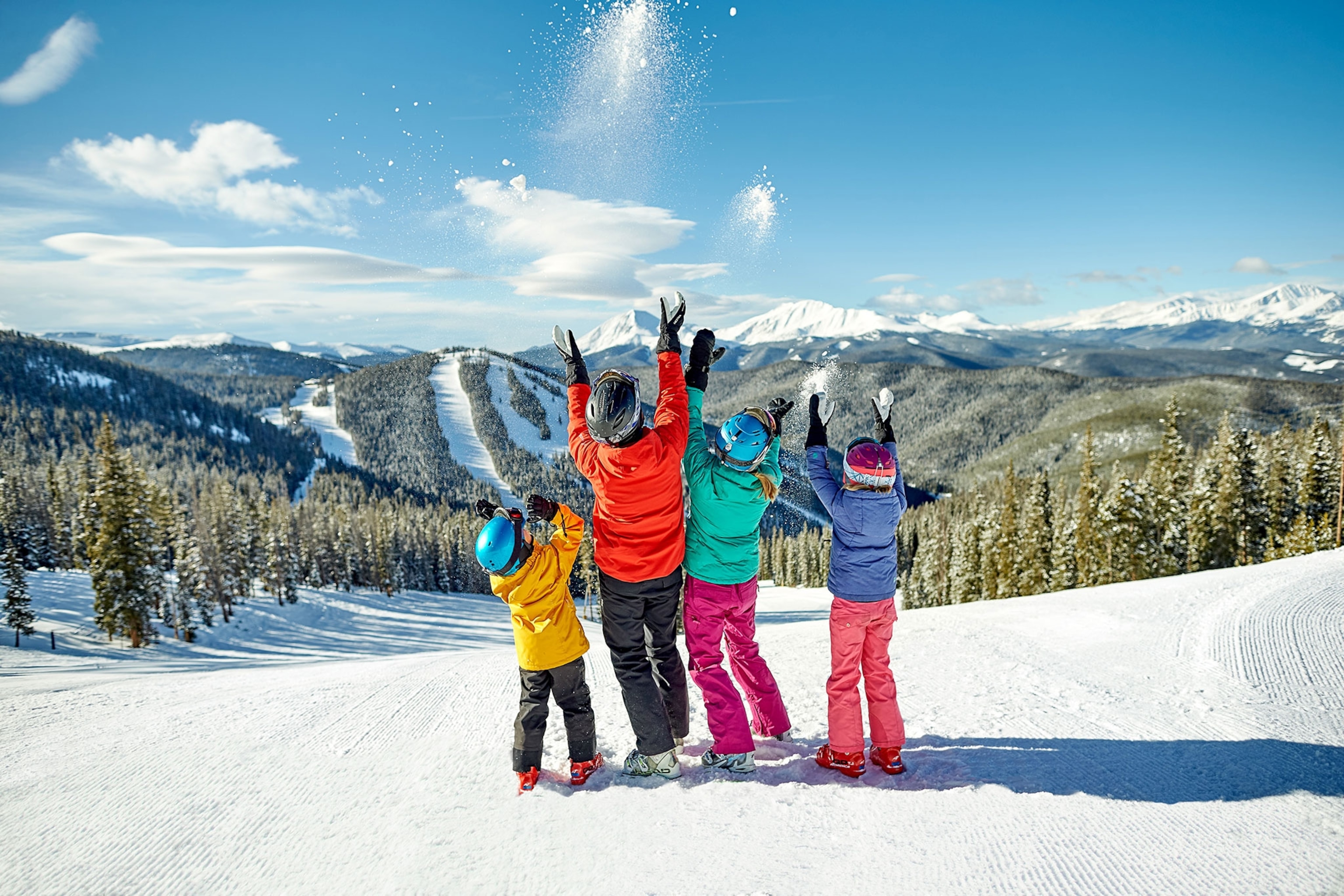 Four people, children and adults, stand at the top of a snowy mountain throwing snow in the air.