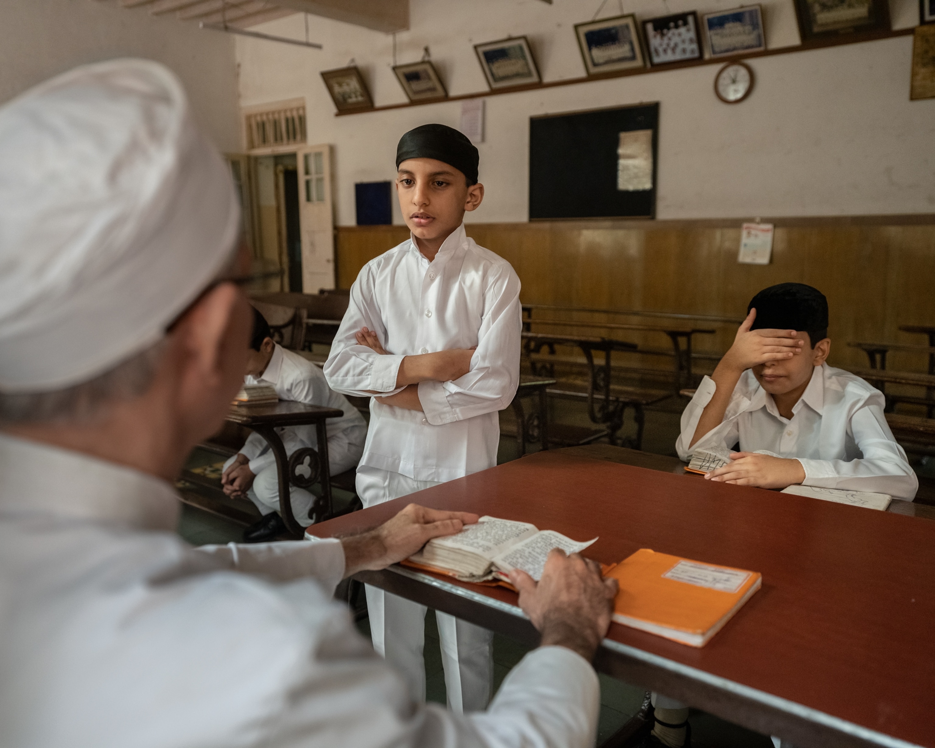 Two young boys (one standing with his arms crossed, one seated with his hand over eyes) are across the table learning from an older male with a book open. The boys are in white clothes and black caps.
