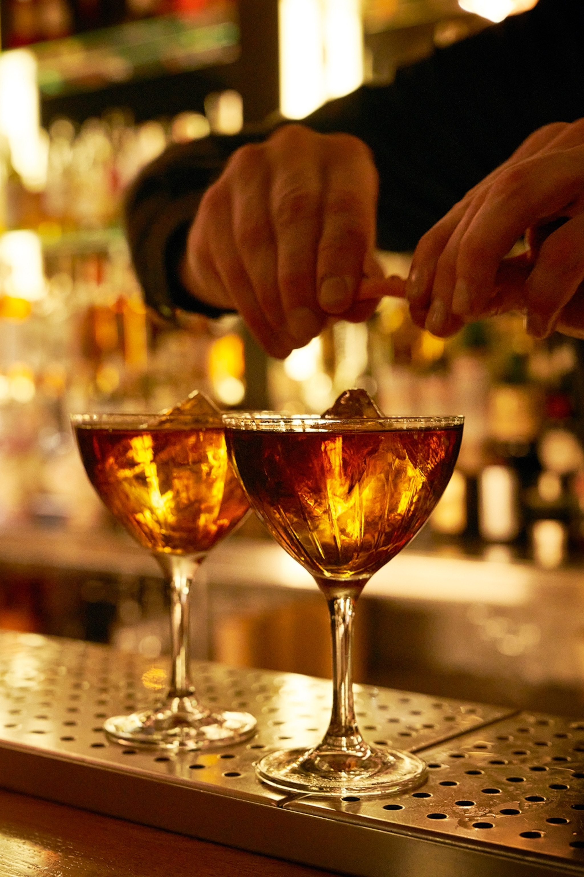 A bartender adding a citrus twist onto two cocktails in a warm, but dart-lit bar in Barcalona.