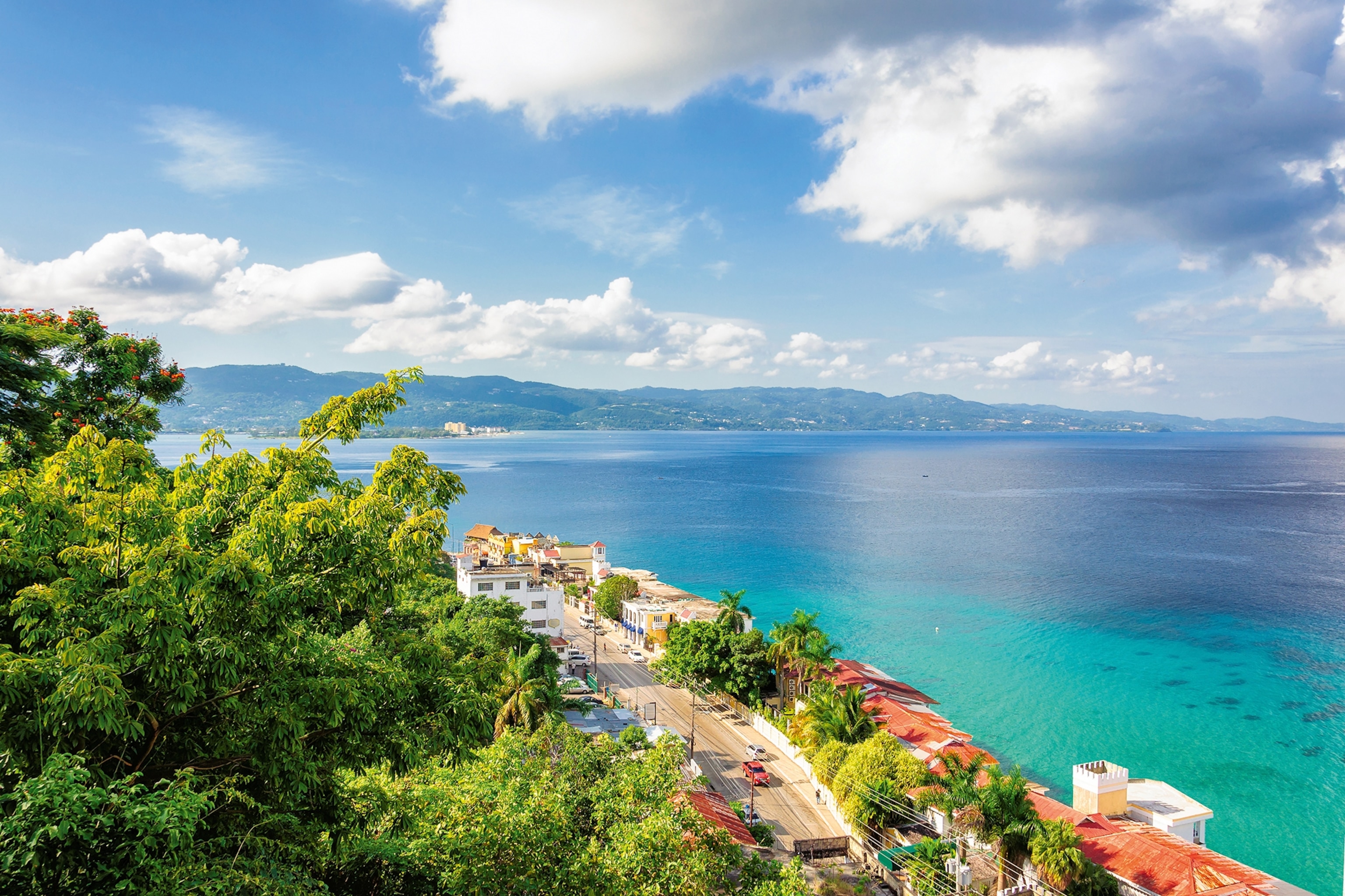 Elevated view of a road lined with lush vegetation and red-roofed buildings running alongside turquoise sea