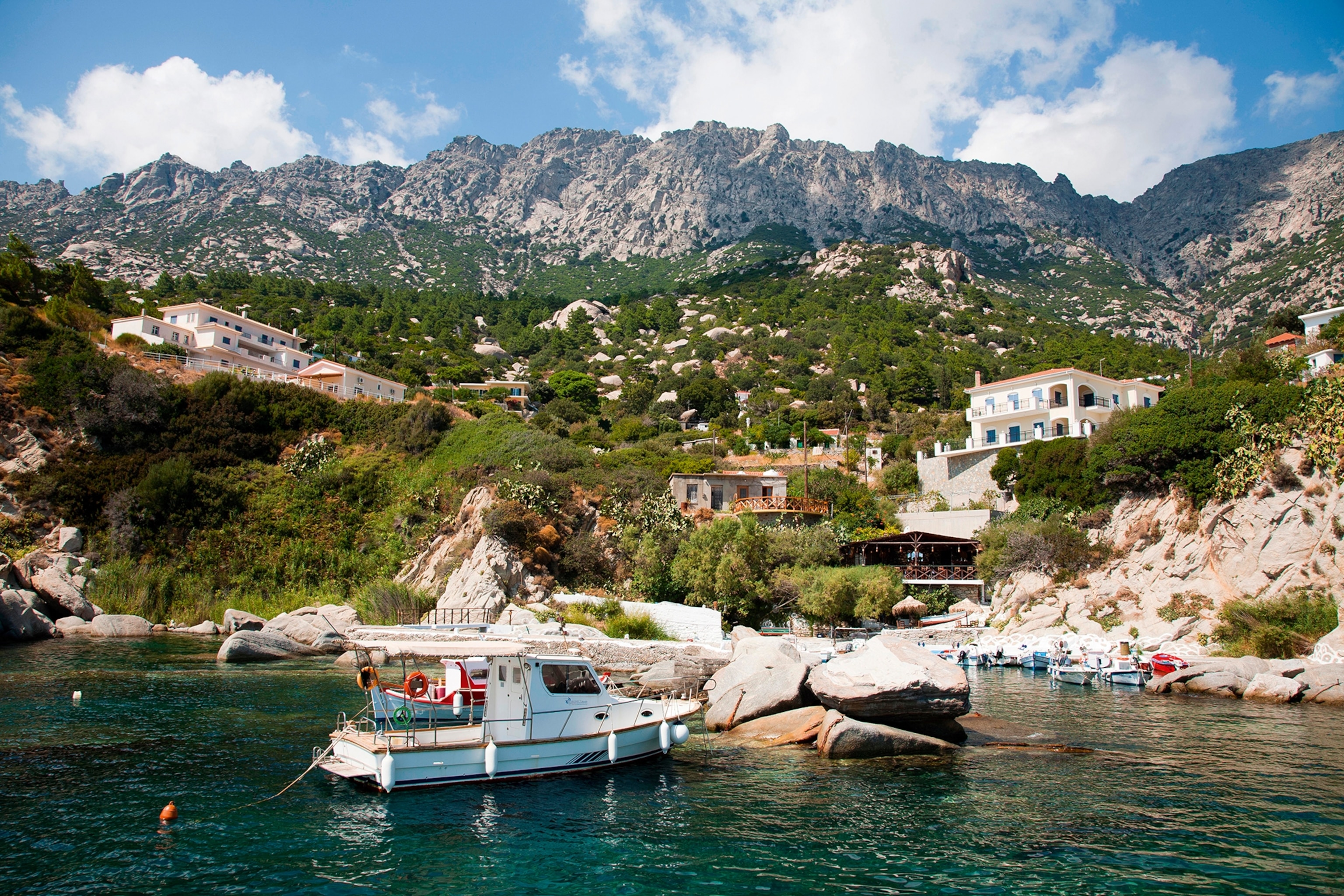 A stone-specked, green-carpeted coastline with mountains in the distance and a small boat in the sea.