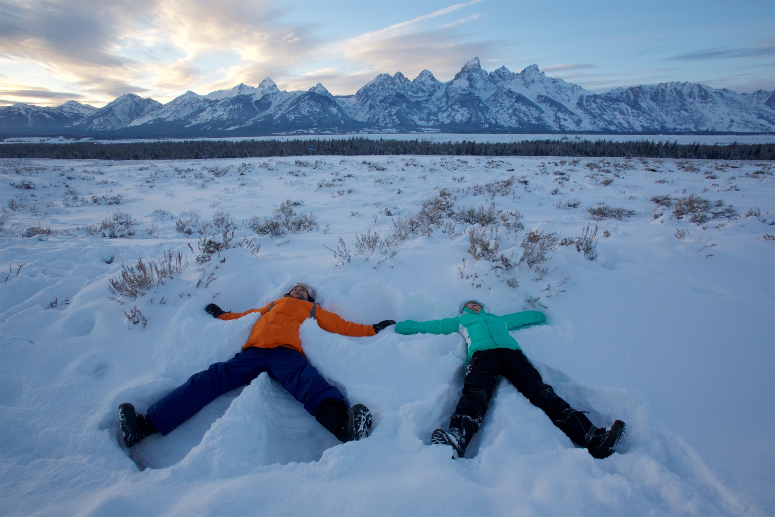 A boy and girl laying in the snow making snow angels with snow-covered mountains filling the horizon in the distance.