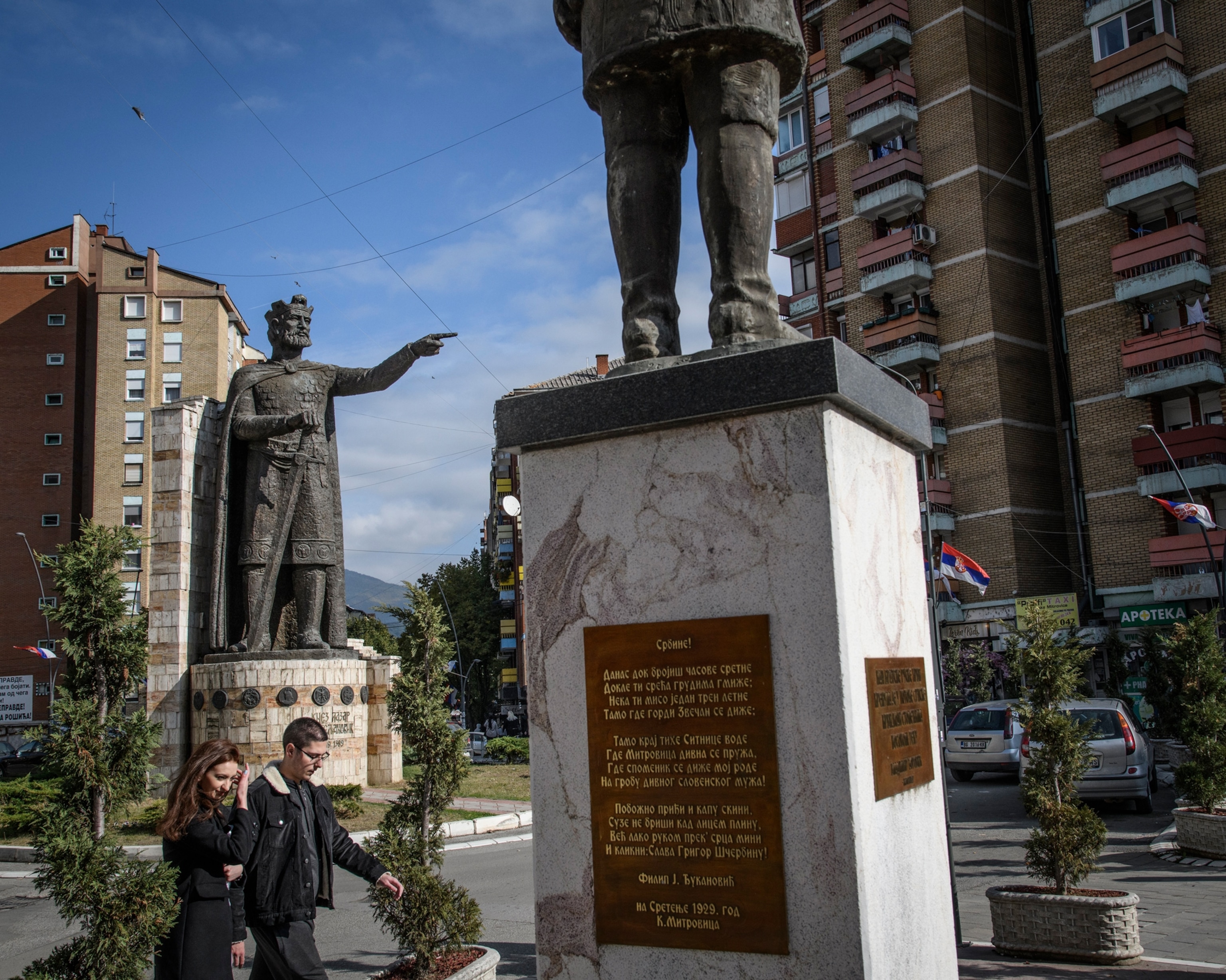 Picture of a large statue of a Kosovo Liberation Army Fighter holding a gun in a city square with people walking below his feet.