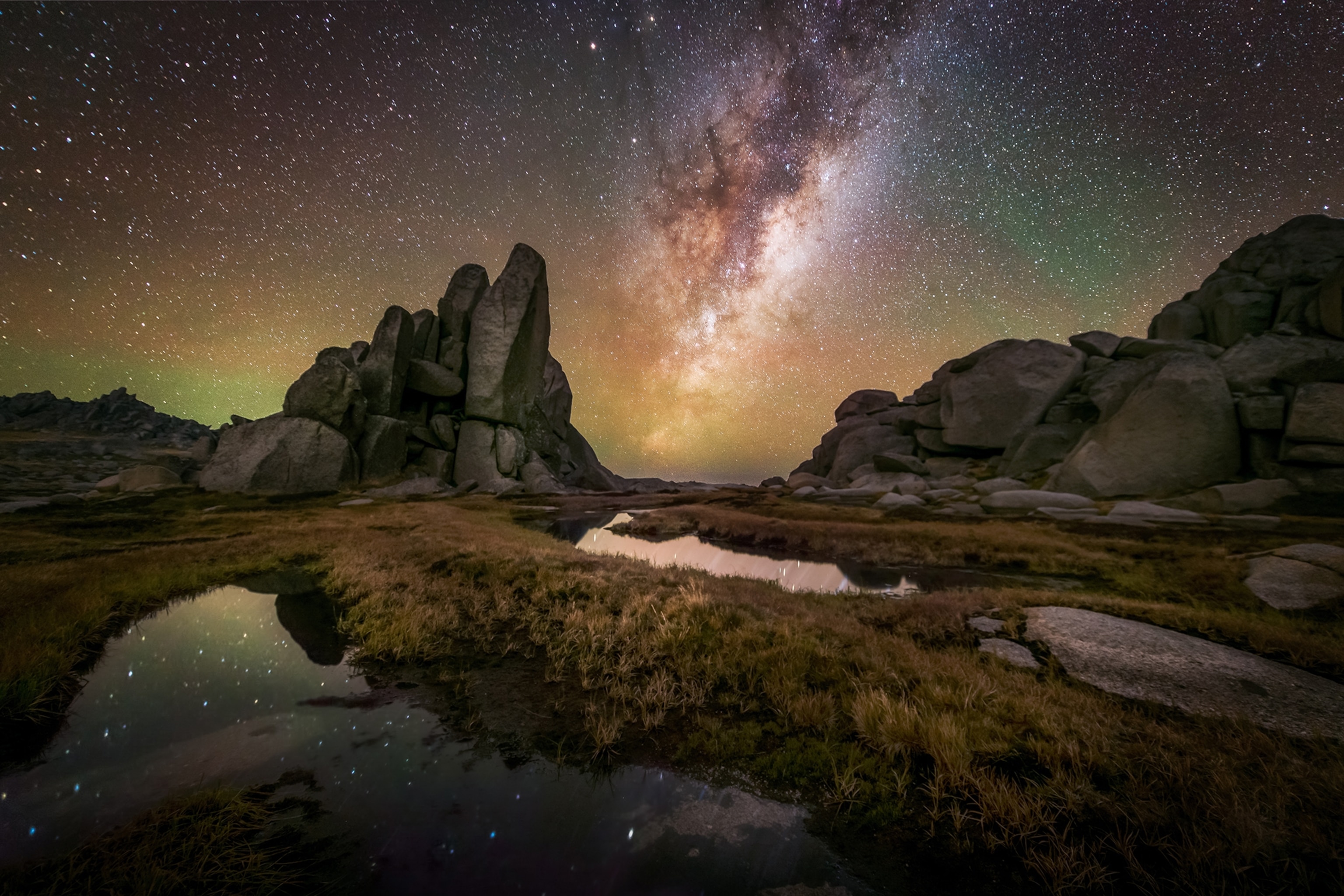 A wide landscape shot at night of an open flat terrain with small ponds and stone formations in the background.