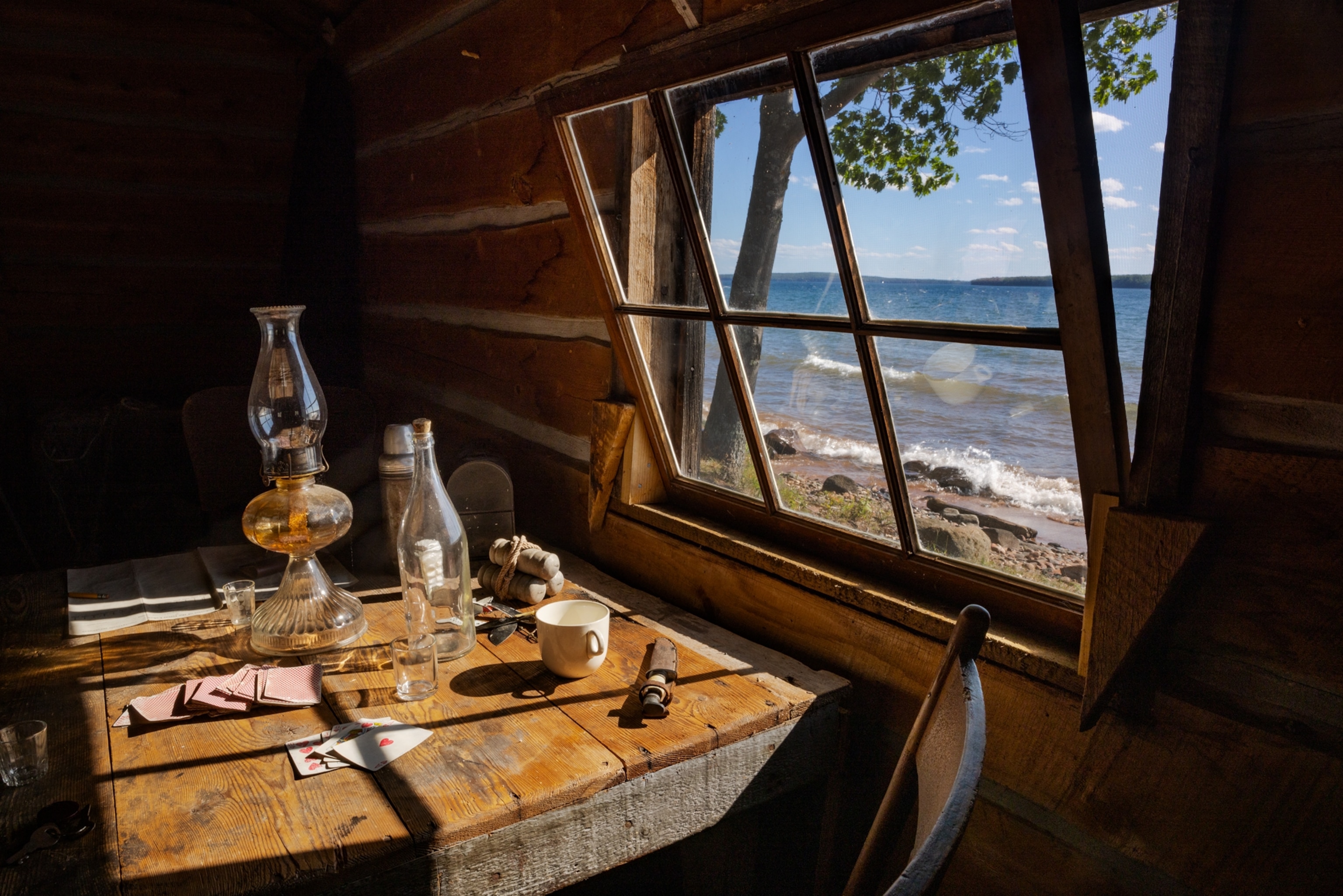 Picture of oil lamp, playing cards, bottle, knife and other small things on the wooden table by the open window looking at the waters.