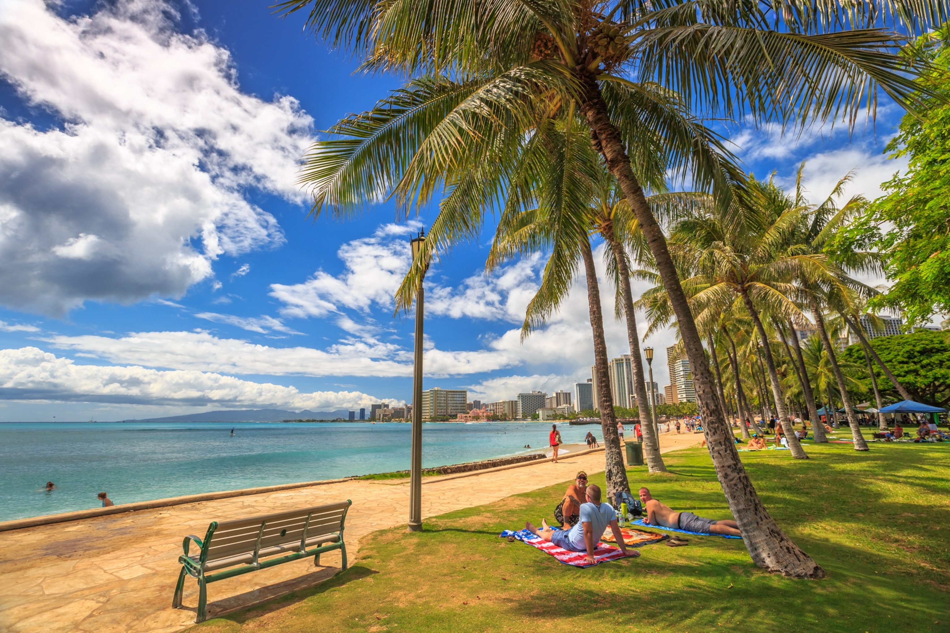 people take the sun lying on the lawn fronting the popular Queen's Beach section of Waikiki Beach. Wooden benches on Queens Beach boardwalk.
