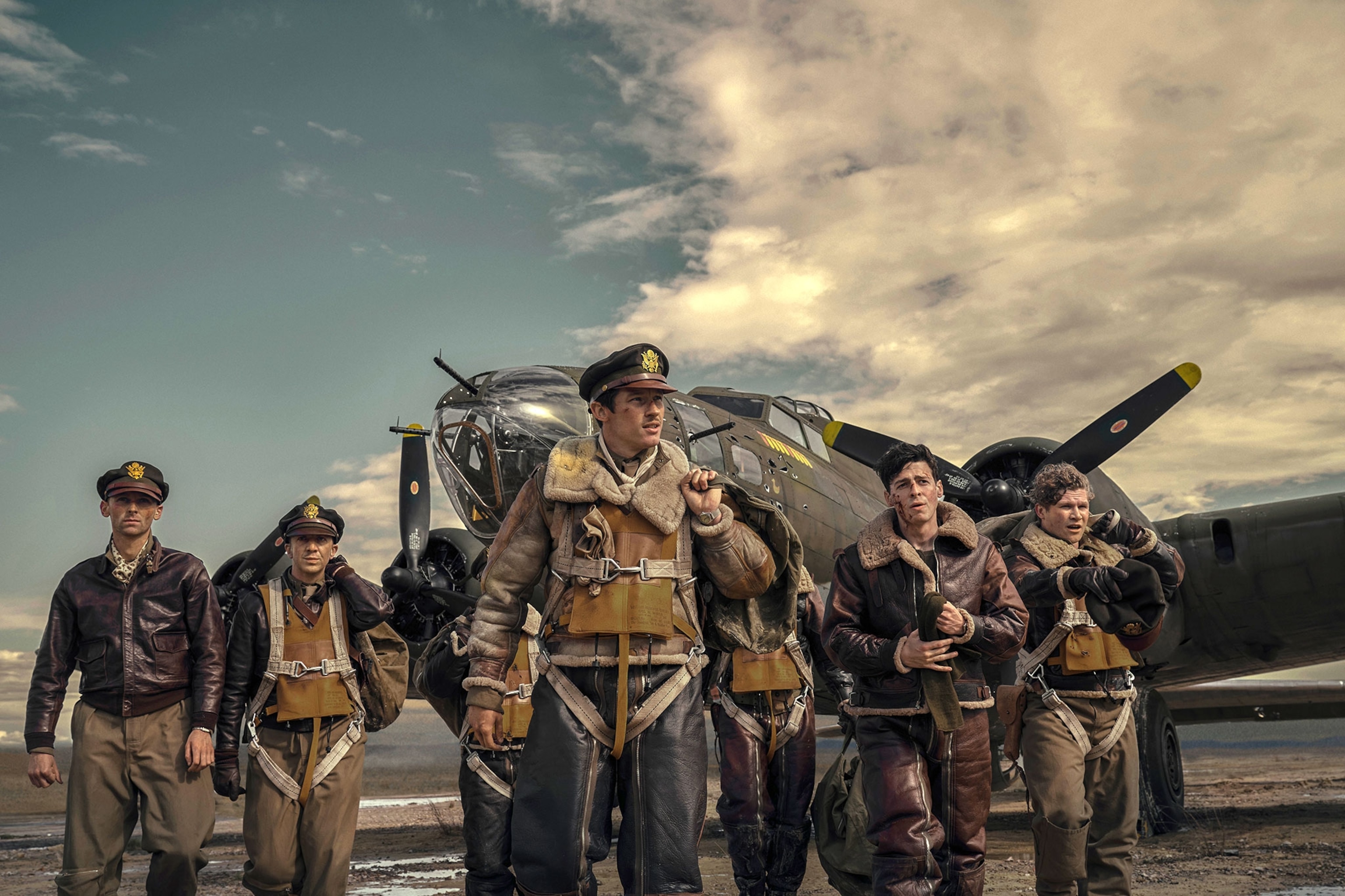 A group of young men, walk away from a military green fighter plane with propeller engines. The men are wearing leather jackes, brown, khaki, and black pants, and leather bomber jackets of different shades of brown. Some men are carrying small canvas duffle bags.