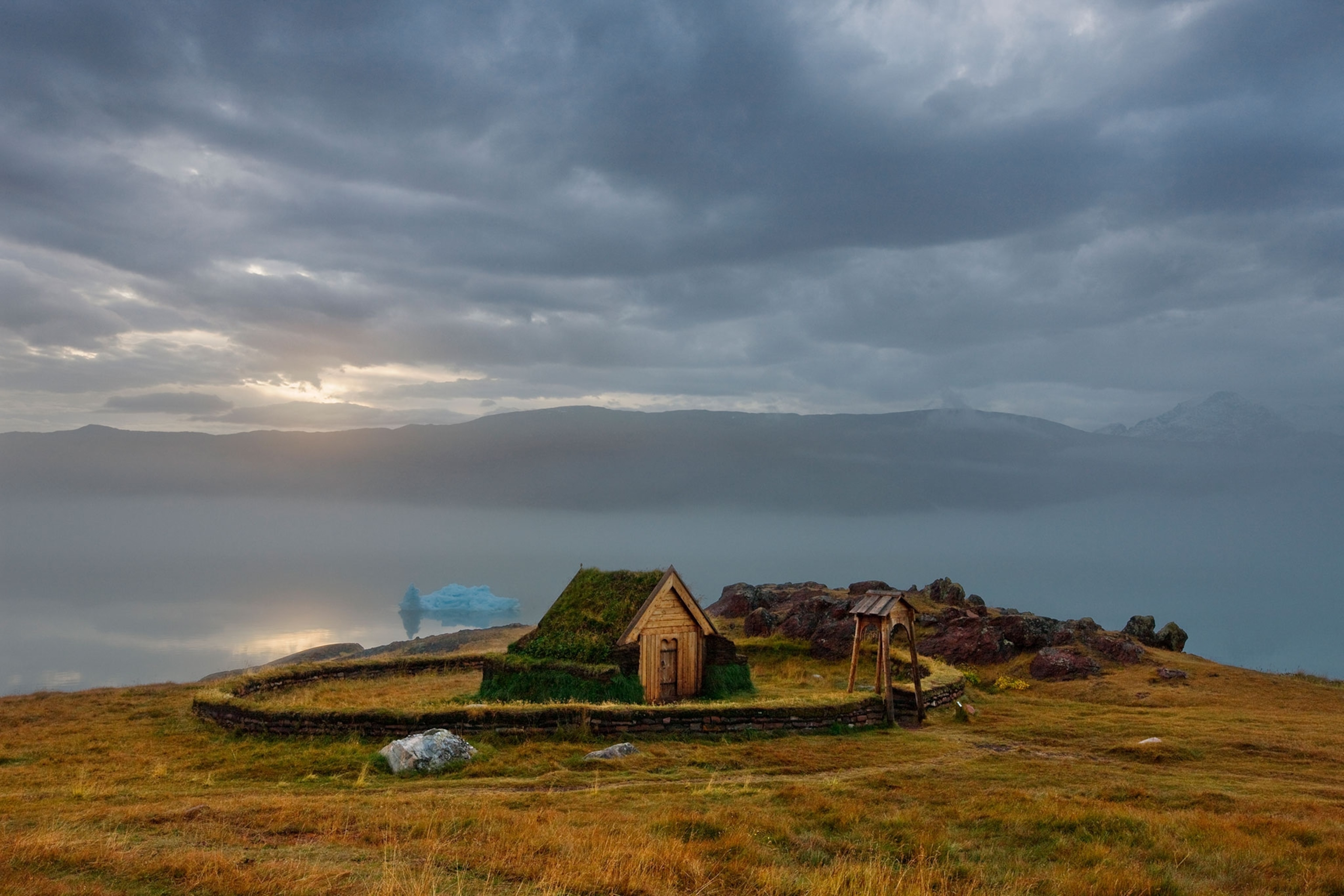 A replica of Erik the Red's wooden church surrounded by a stone wall sits near the edge of a cliff. The hill is covered in yellow grass and big rocks with water and mountains peaking through fog in the background.