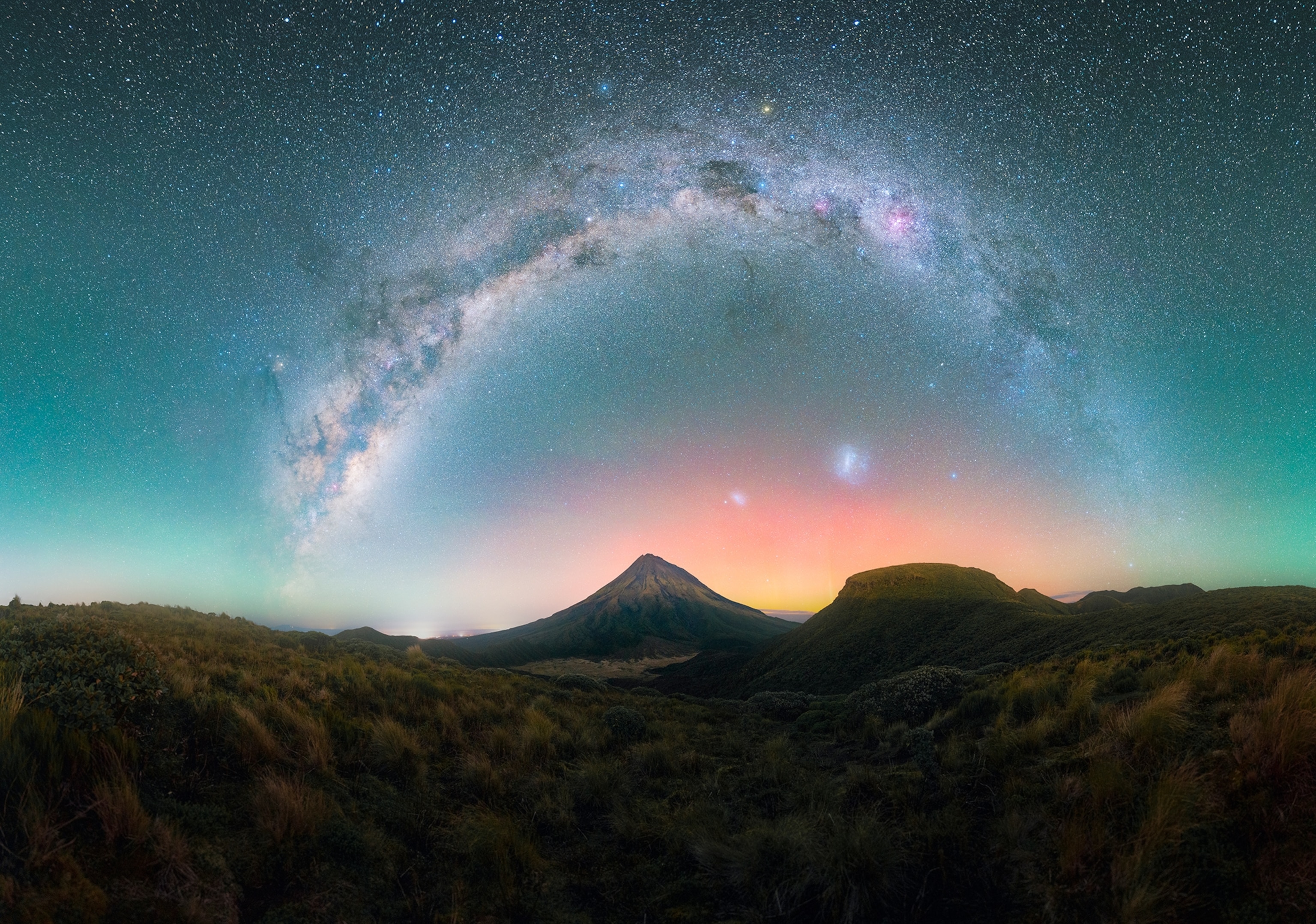 Under the sweeping arc of the Milky Way, Mount Taranaki stands sentinel in Egmont National Park, New Zealand, silhouetted against the faint glow of the Aurora Australis on the horizon.