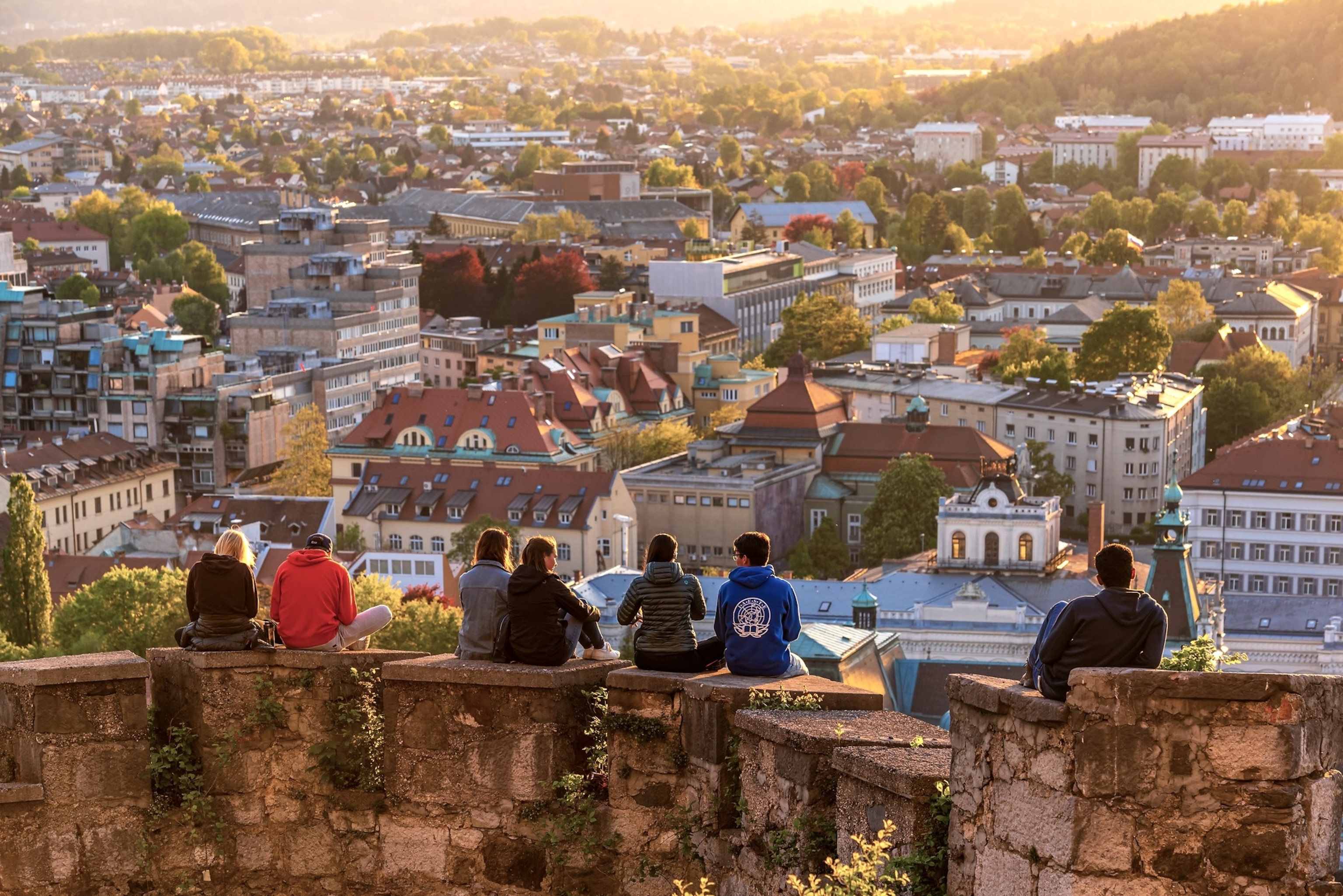 The backs of seven men and women sitting on the edge of a stone wall look high over rooftops of a city.