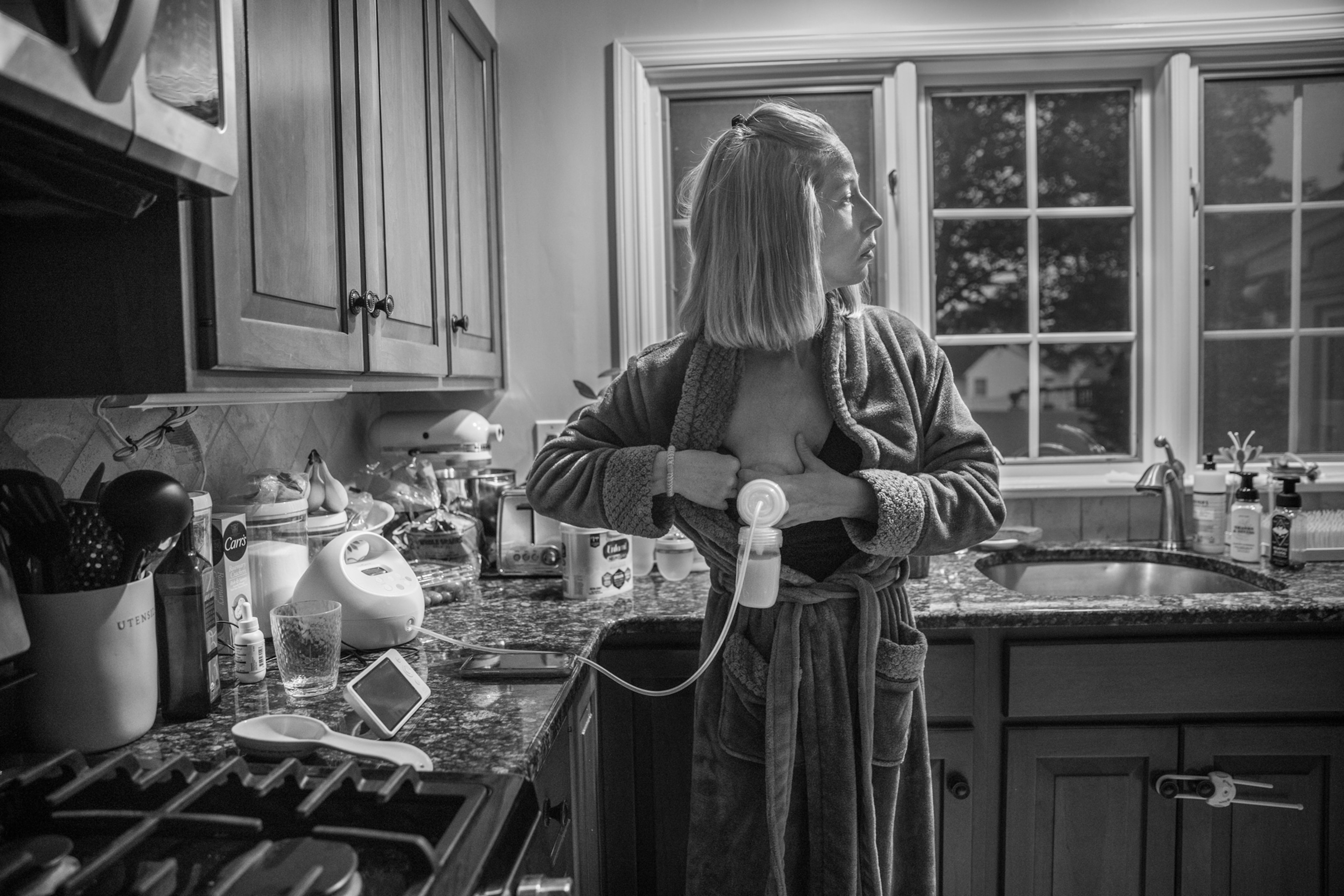 Woman pumping breast milk in the kitchen and trying to see what is happening in another room.