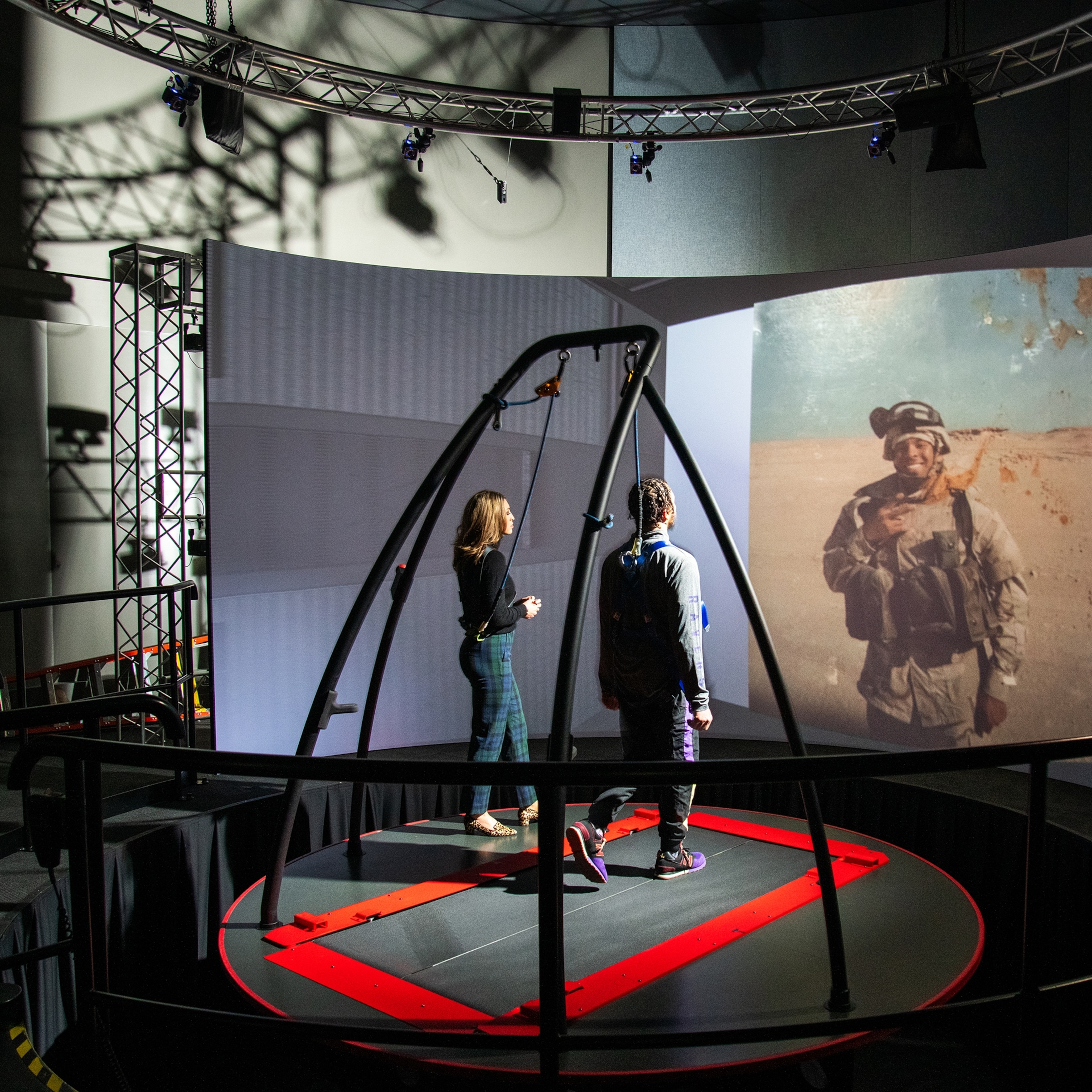 A man wearing a harness walks on a treadmill apparatus towards an old photograph of himself as a war soldier projected on the screen in front of him. A woman stands on his left for support.