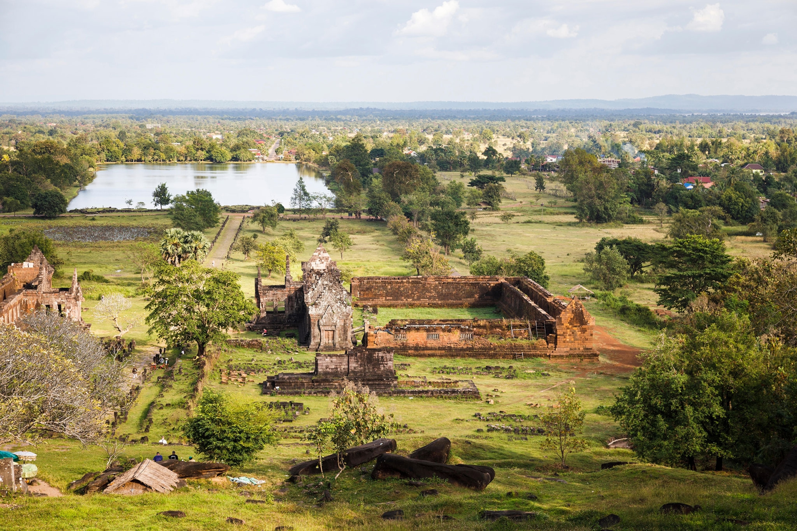An aerial view of ruins, green jungle, and a town far off in the distance with blue sky and clouds.