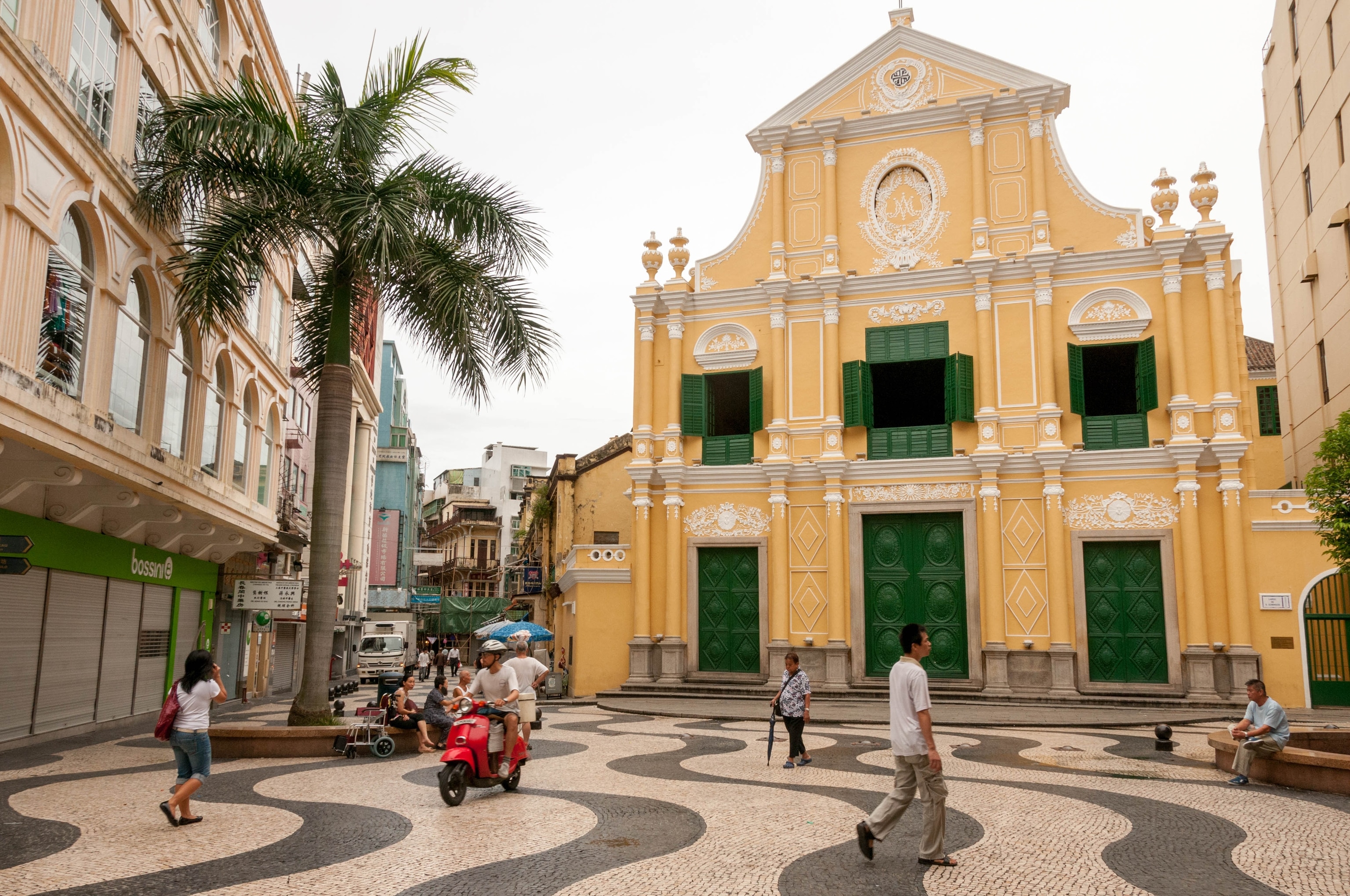 Floors a paved in a rippled black and white pattern, a yellow church with green doors fill the frame as people travel through the area.