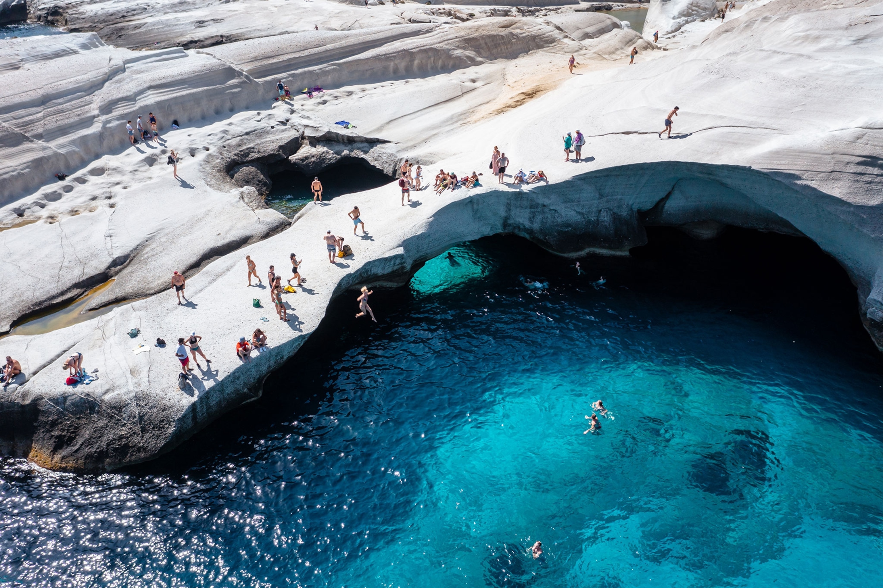 Aerial view looking down at people jumping from a big rock covering the top half of the frame into blue water.