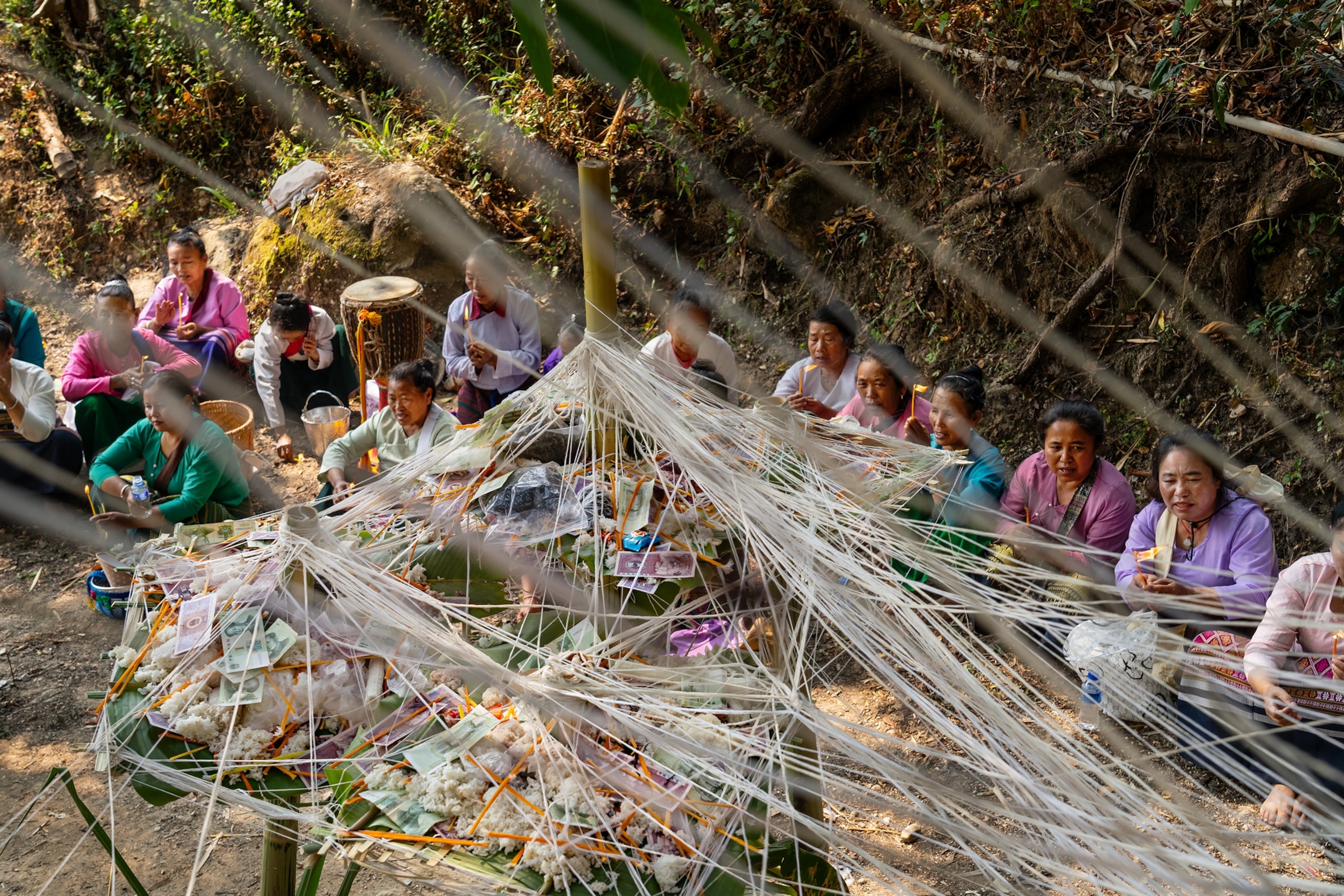 A group of women sit around a bamboo post that carries various symbolic objects. White strings form a net around the object and stream through the photo from one end to the other.