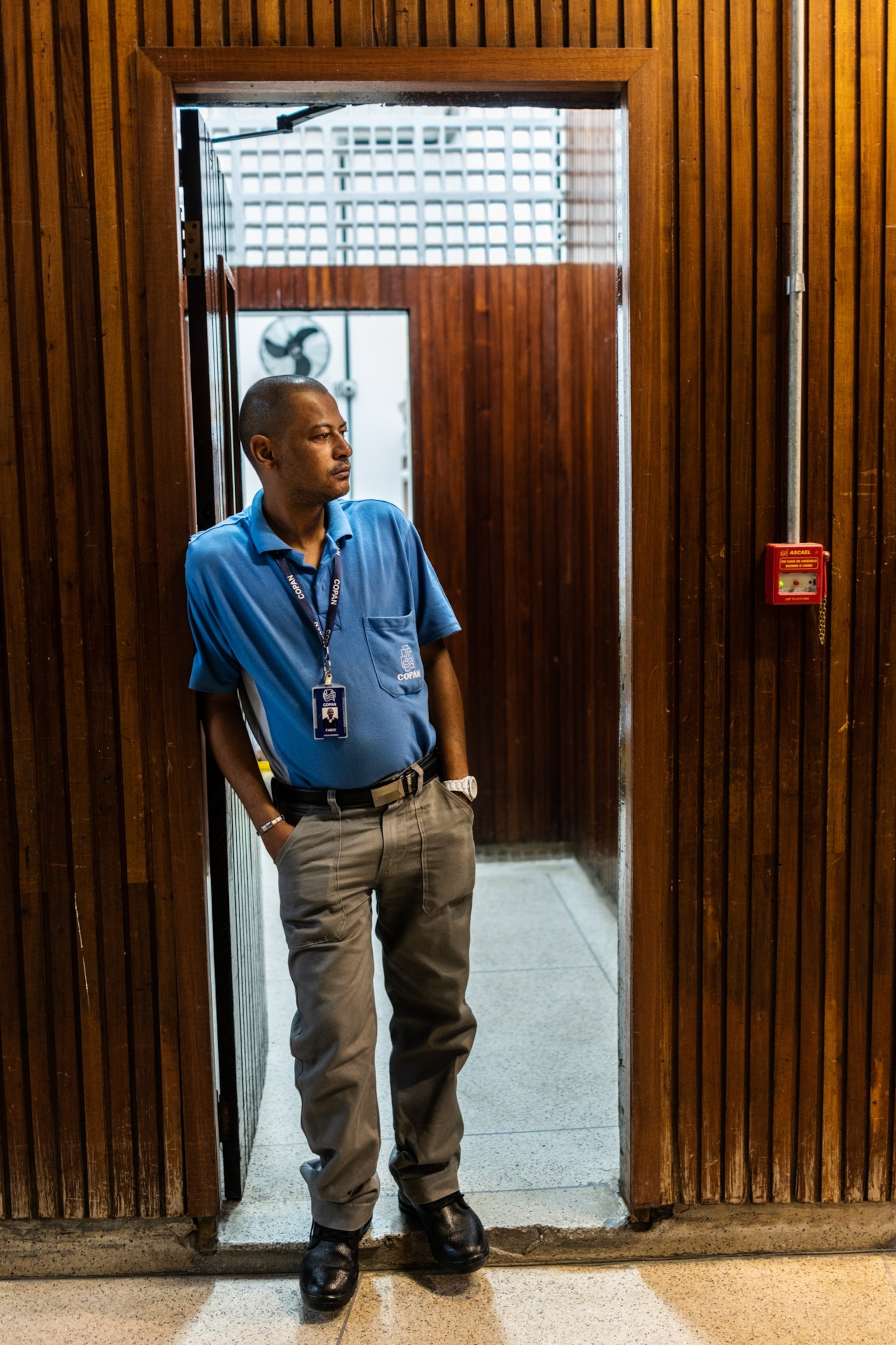 a man in a blue collared shirt leaning against a wood paneled wall