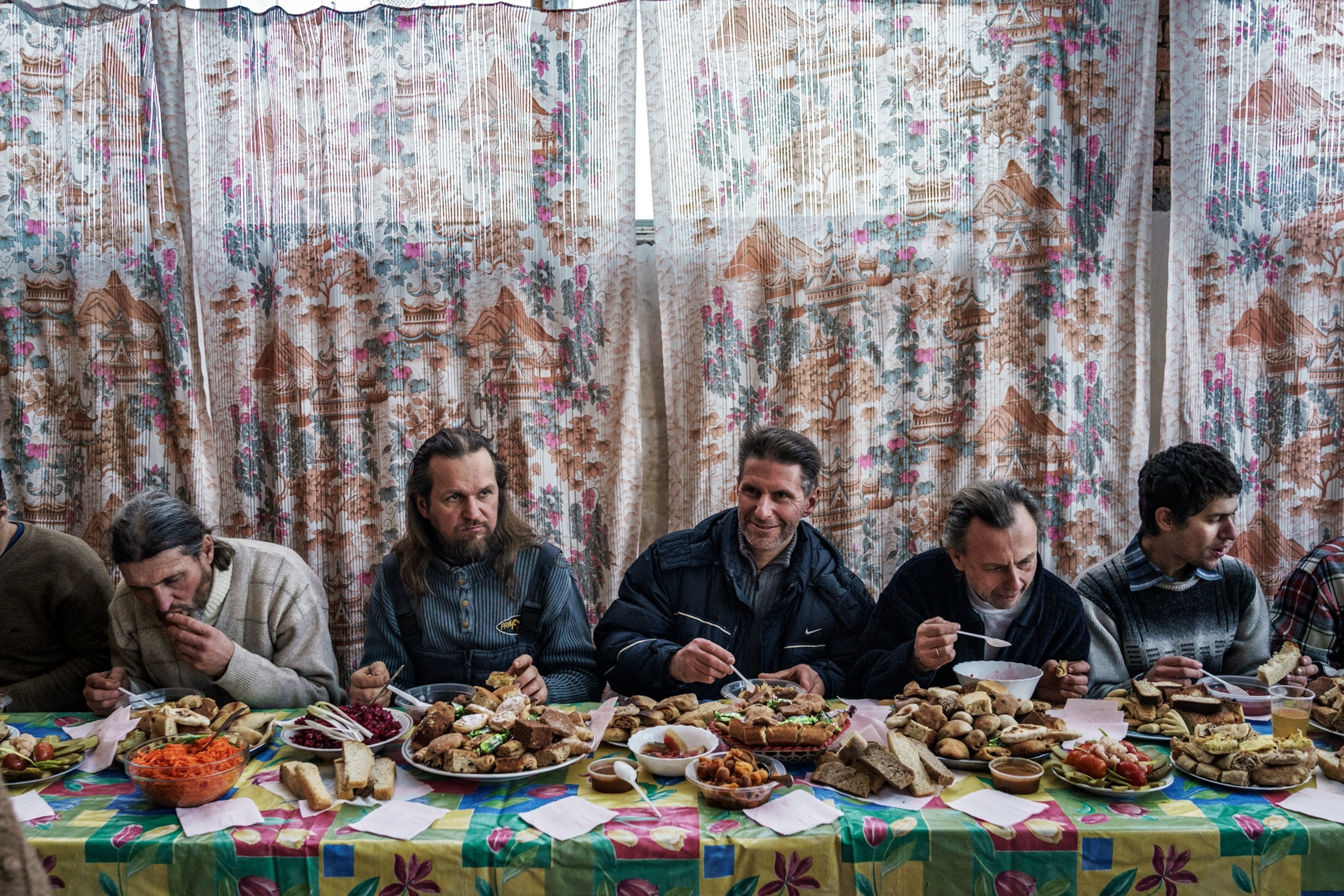 men sitting on one side of a table feasting on lots of food on a colorful tablecloth