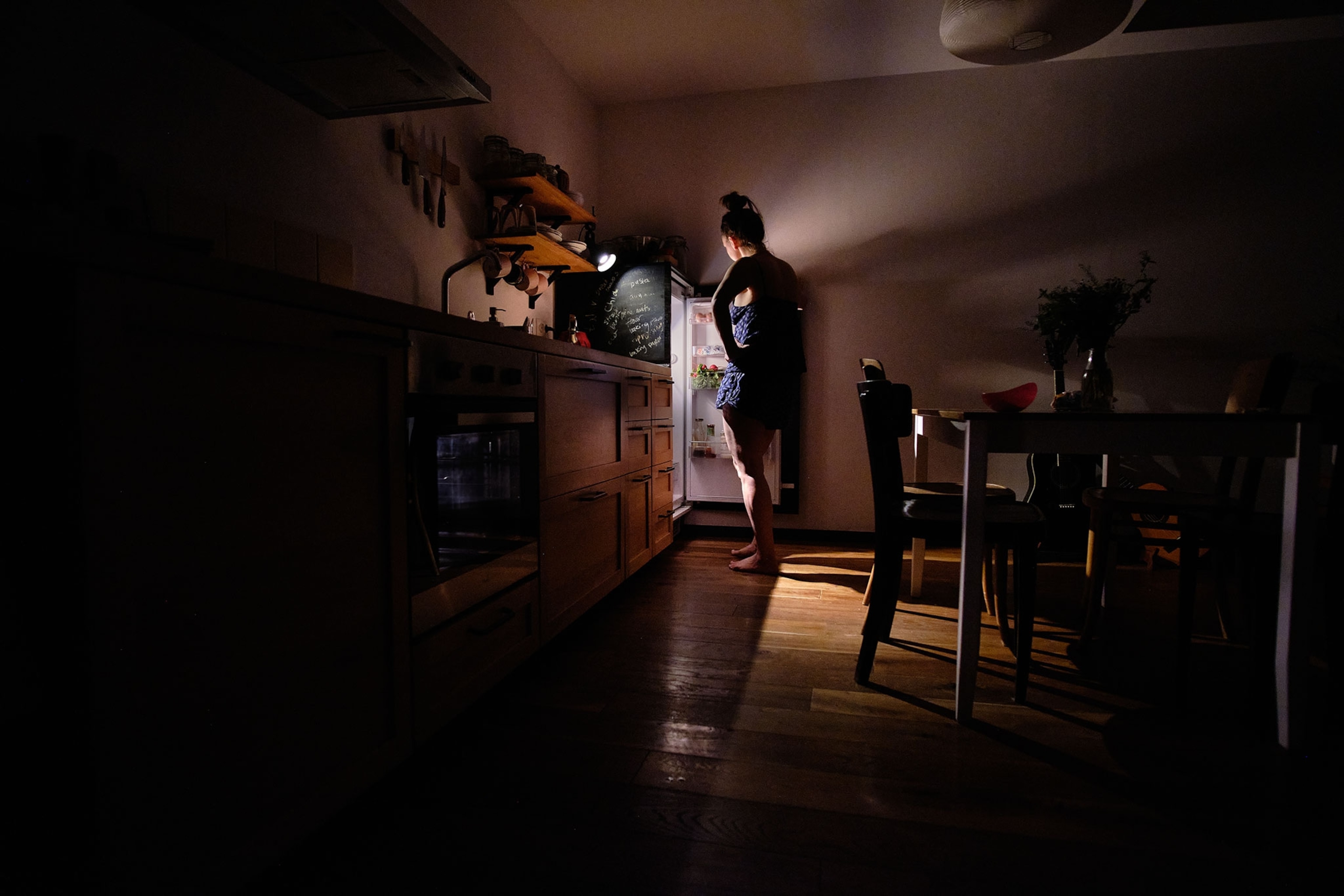 A woman stands in a dark kitchen peering into a fridge that sits in the corner, the light illuminating her and the kitchen.