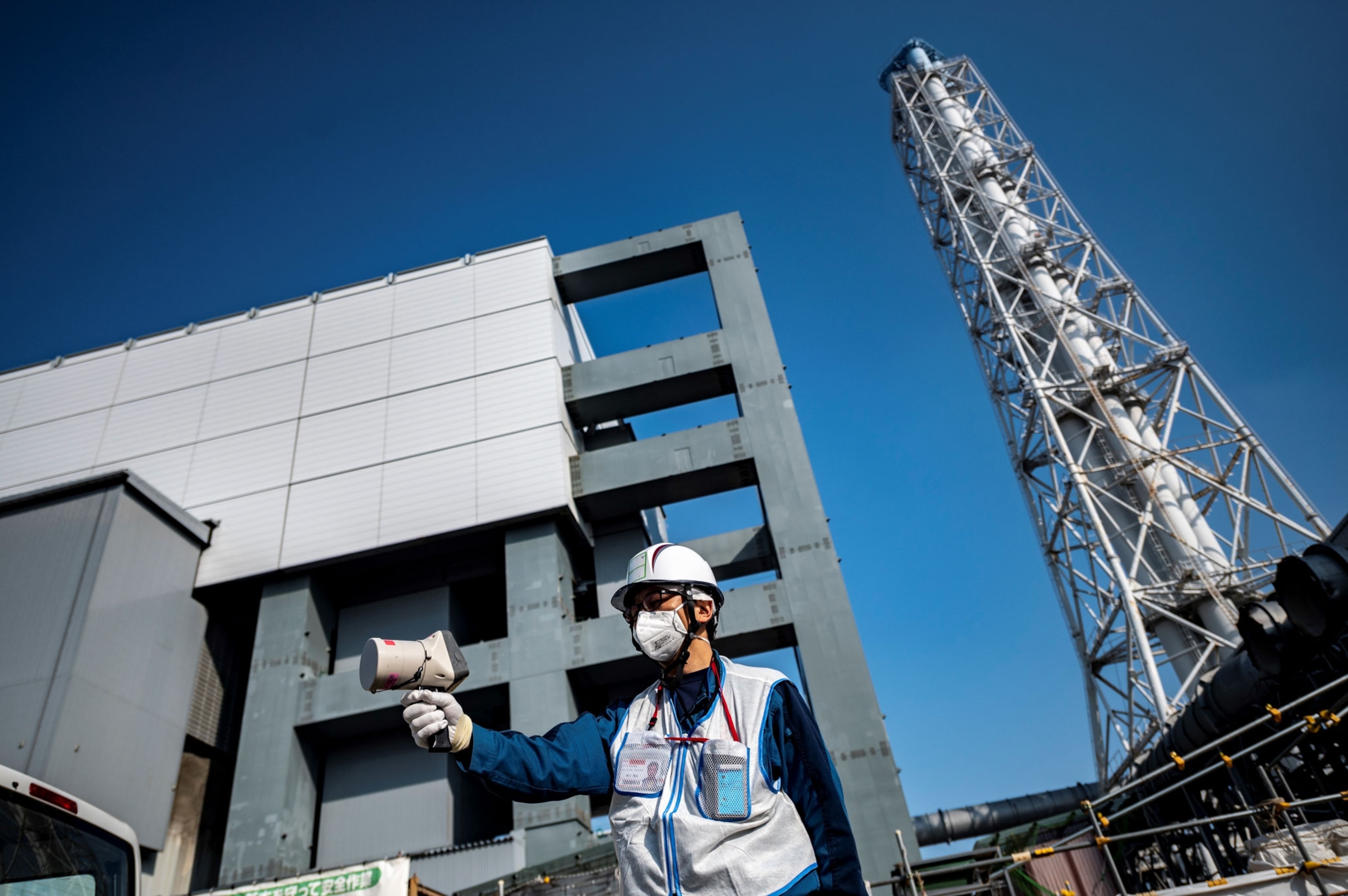 a person in a mask and a hard hat holds a geiger counter to measure radiation in front of the Fukushima nuclear power plant