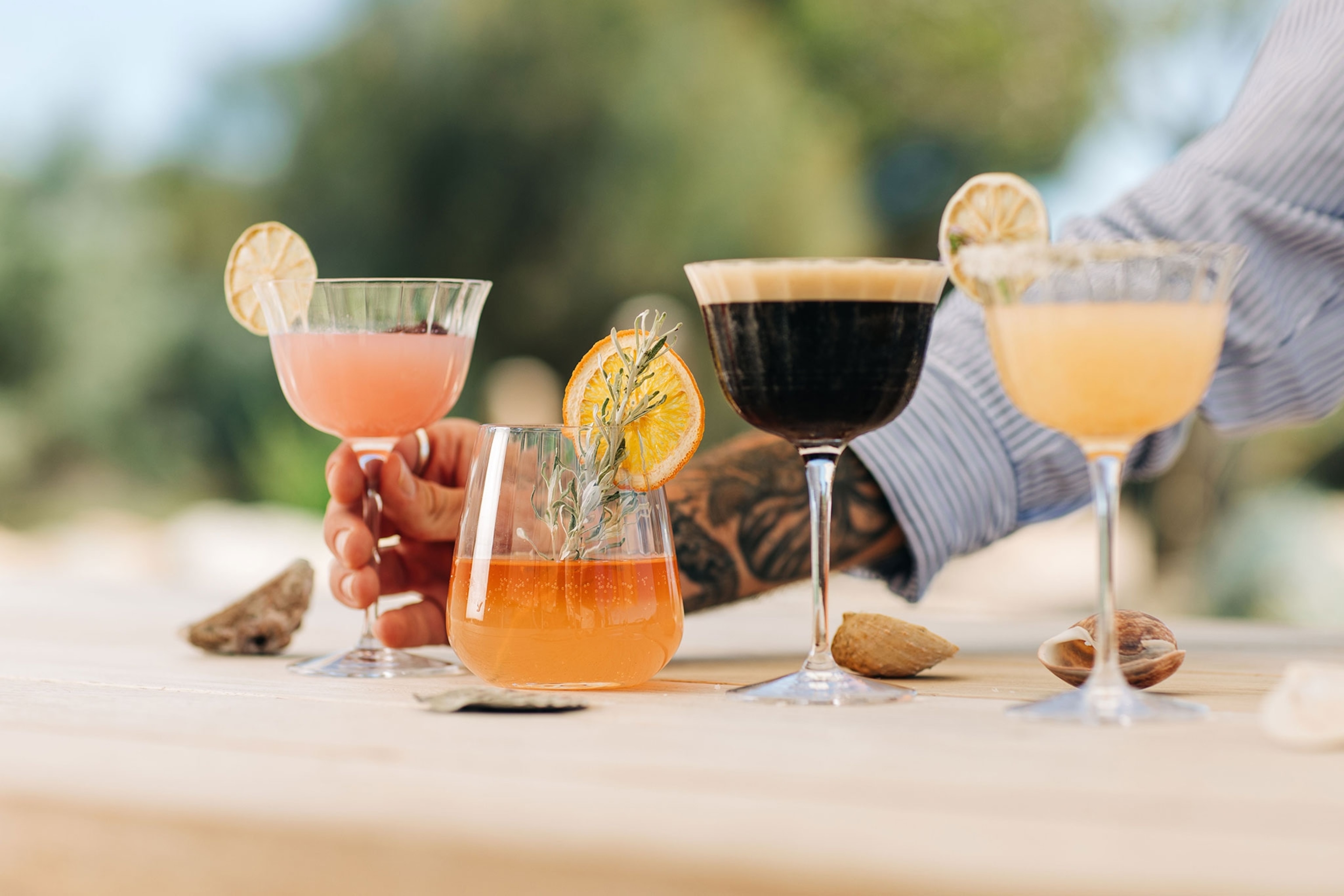 Four unique cocktail drinks lined up on a light wooden surface with an arm reaching for one.