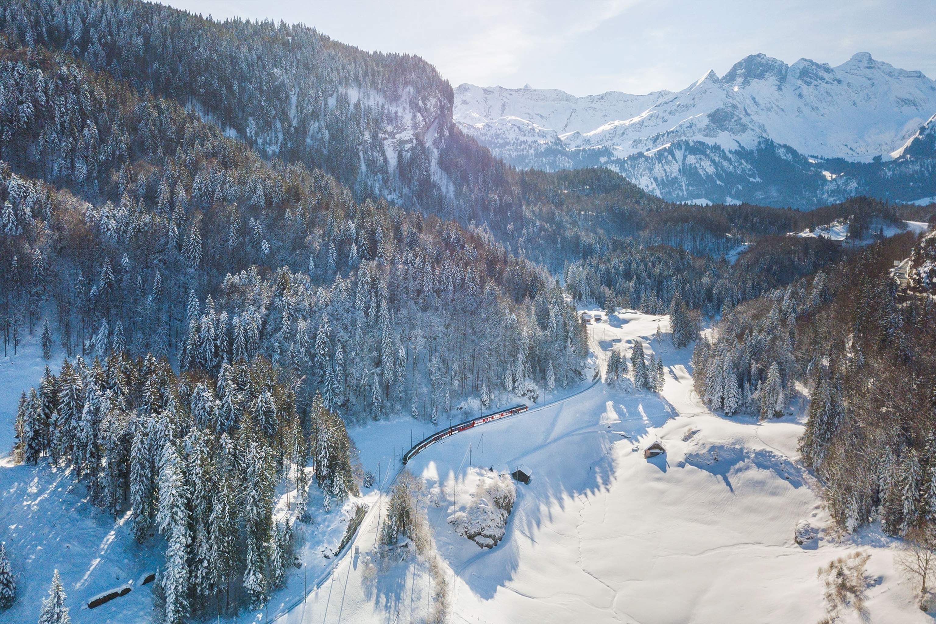 An aerial view of the Brunig Pass shows a train gliding through snow-capped landscapes, with pine trees and mountain peaks in view.