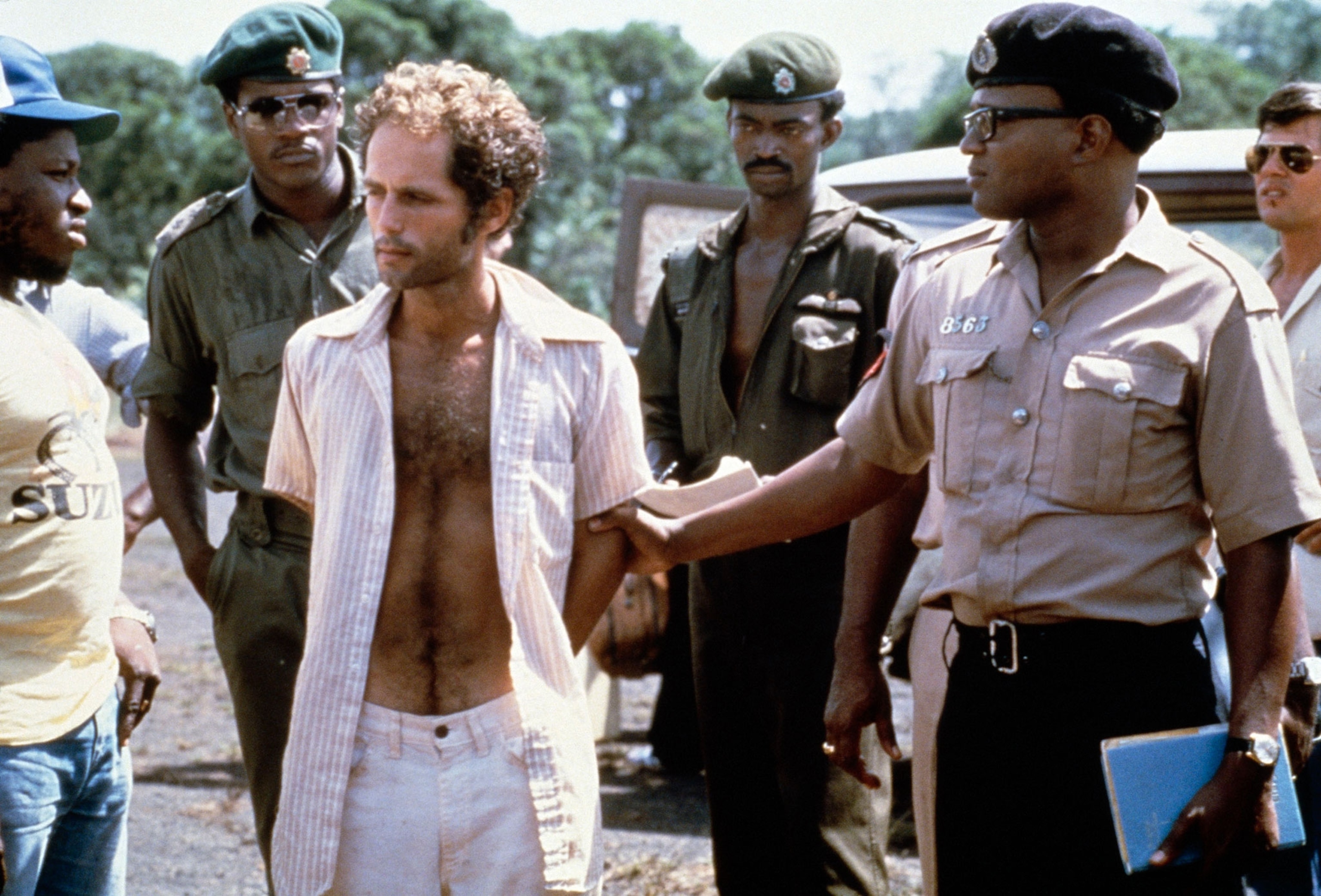 Larry Layton in white paints and a striped button up shirt stands with guyana police in following his arrest